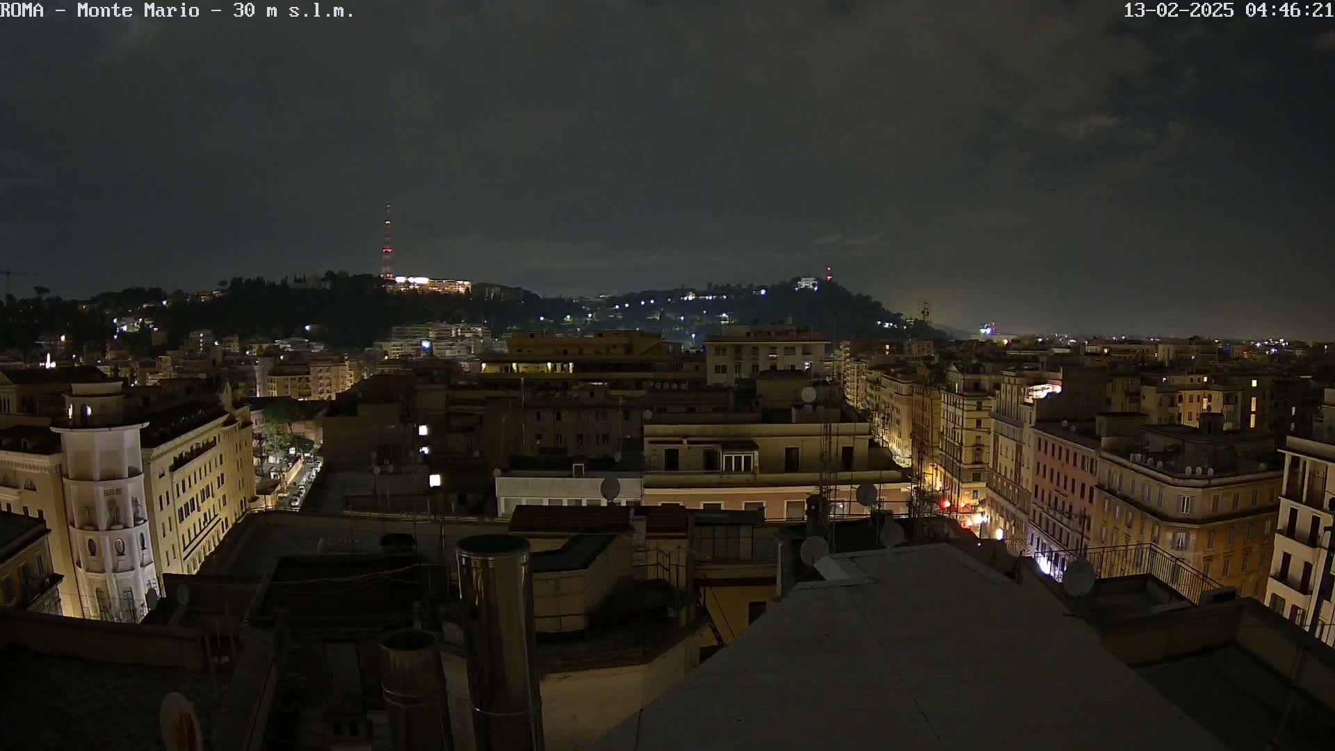 A nighttime cityscape of multi-story buildings, illuminated by streetlights and windows, with a hill in the background under a dark, cloudy sky.