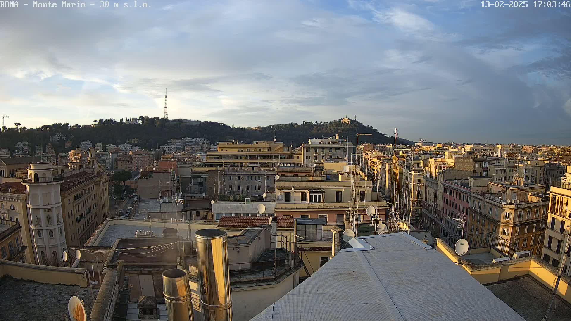 A mostly cloudy sunset illuminates a cityscape of closely packed buildings, with a hill in the background.