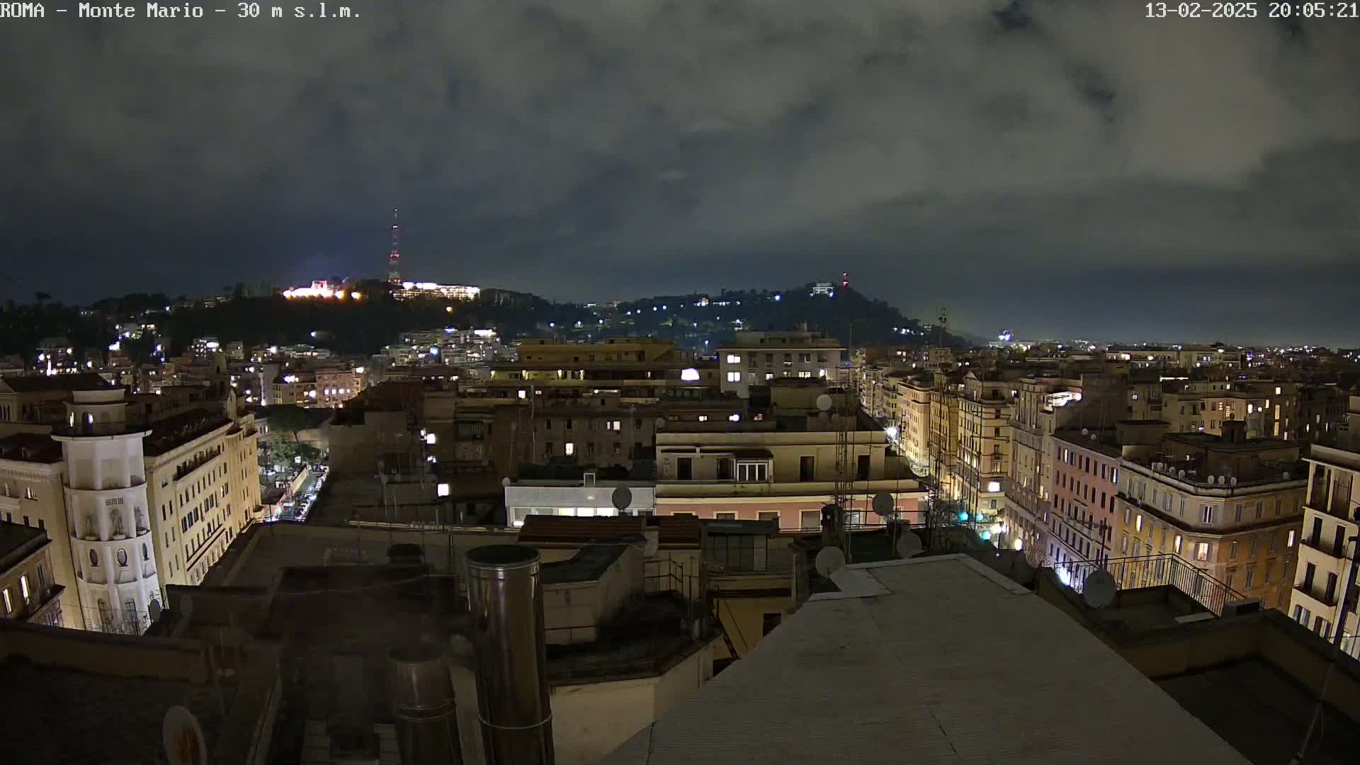A nighttime view of a city illuminated under a cloudy sky, showing a hilltop in the distance.