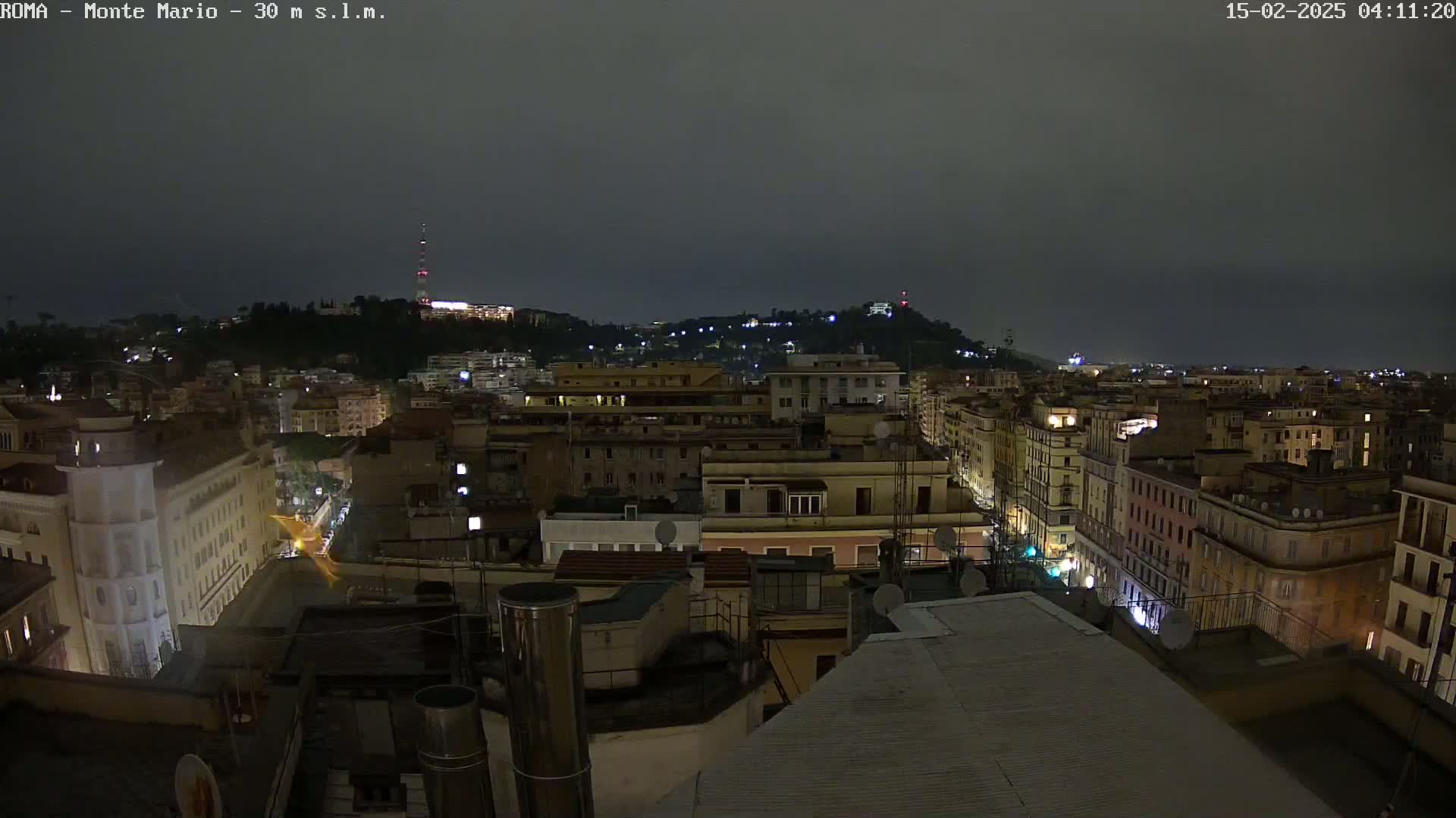 A nighttime view of a city, with hills in the background under a dark, overcast sky.