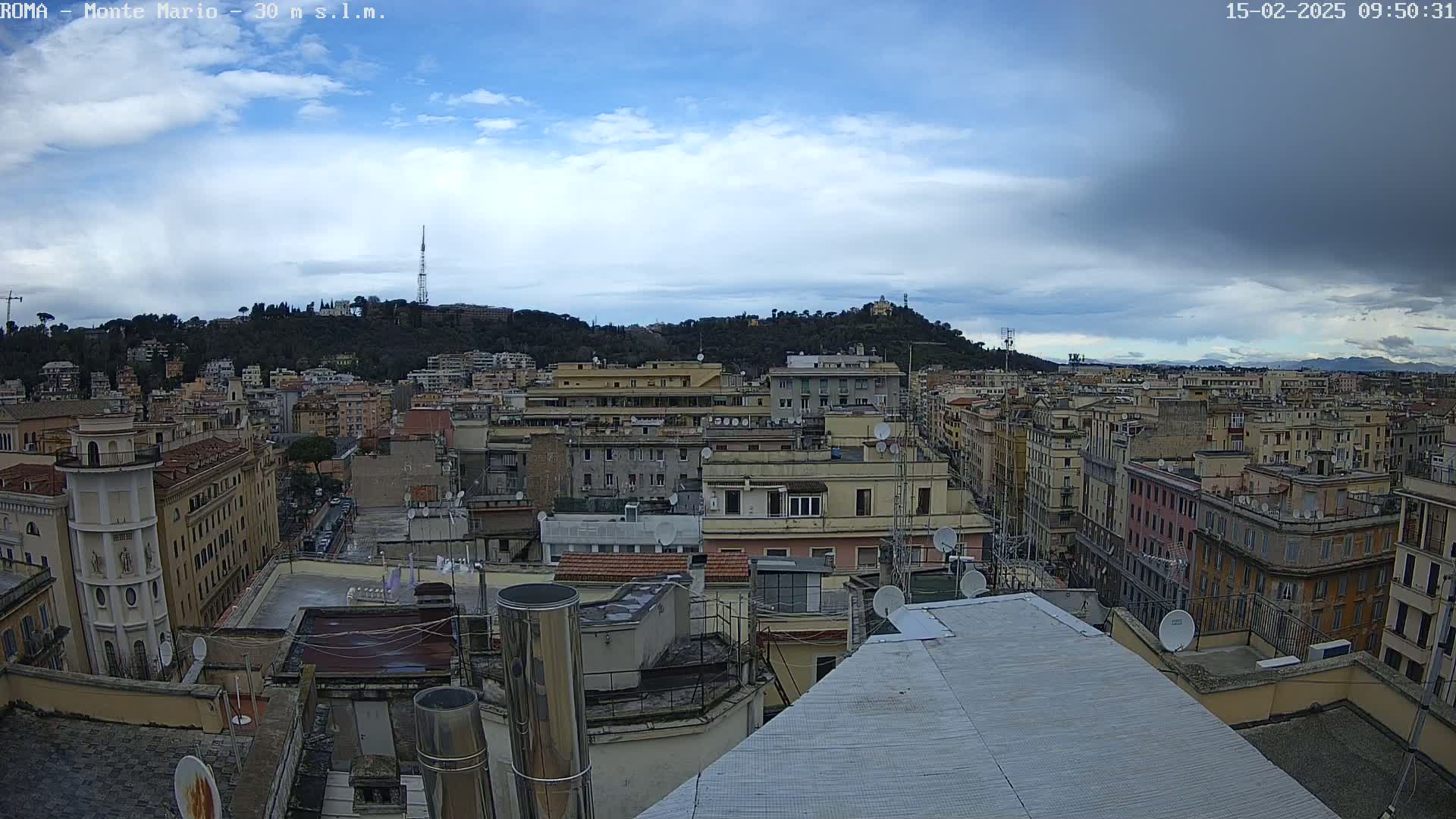 An aerial view of a city under a partly cloudy sky shows many buildings with rooftops and satellite dishes, with a hill in the background.