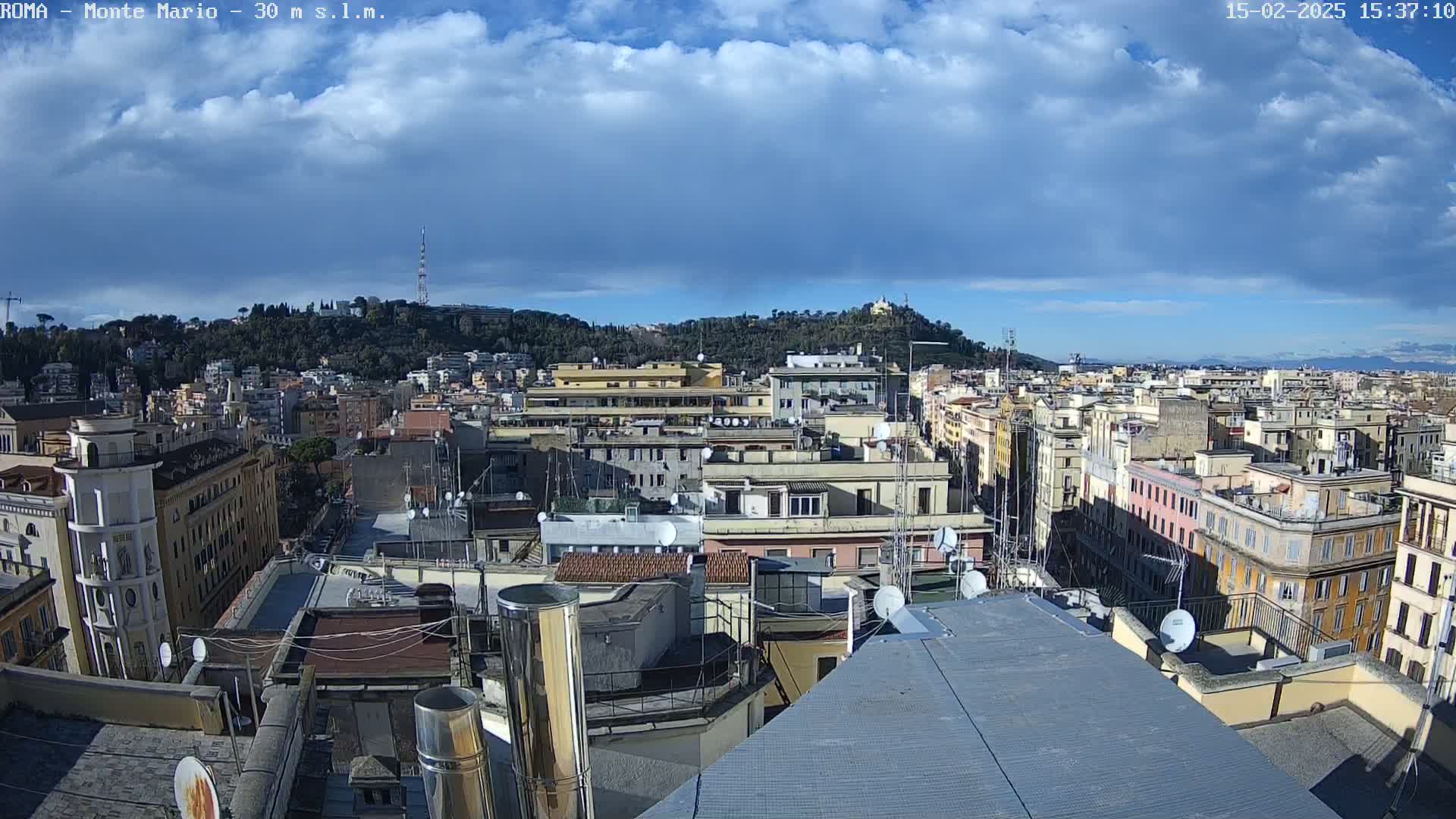 A partly cloudy sky overlooks a densely packed cityscape of multi-story buildings, with a hill covered in trees visible in the background.