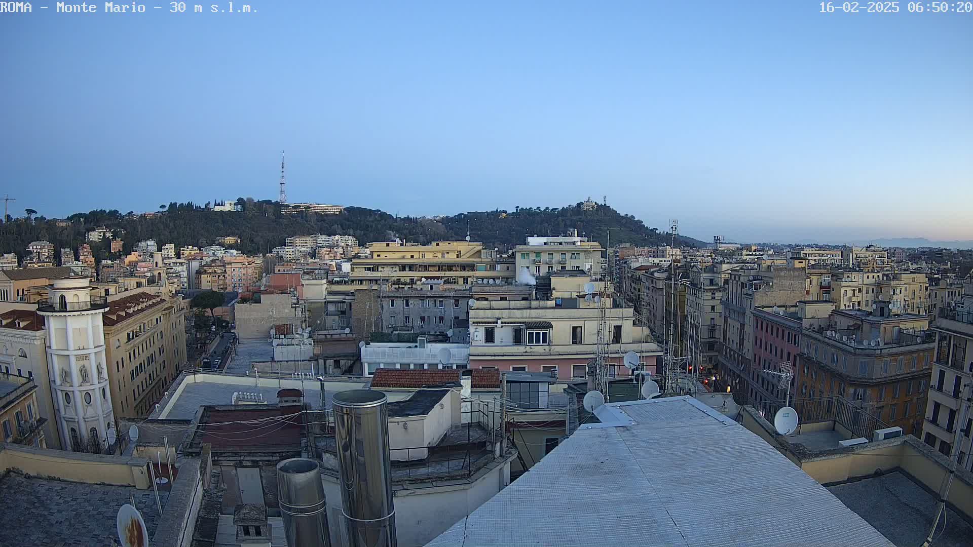 A panoramic rooftop view of a city under a clear, twilight sky, showing numerous buildings extending to a hill in the distance.