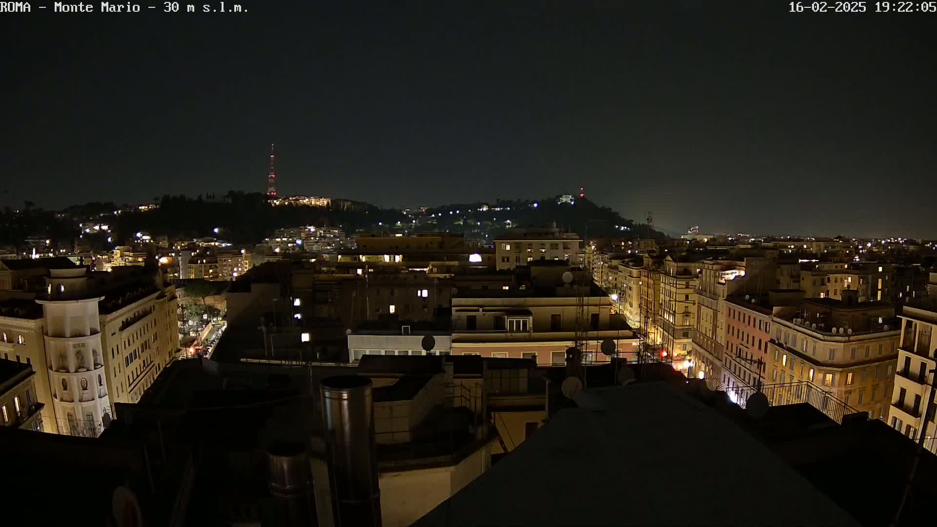 A nighttime view of a city, showing many buildings illuminated, with a hill in the background under a clear, dark sky.
