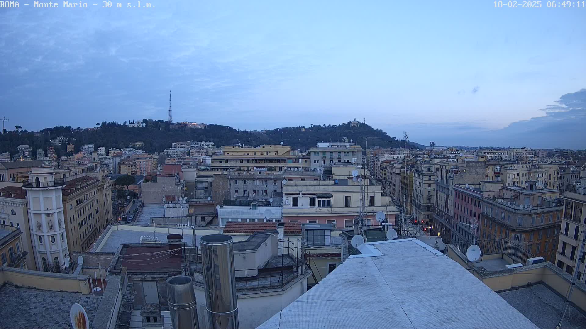 A cityscape at dawn, with a hill in the background under a mostly clear, light blue sky.