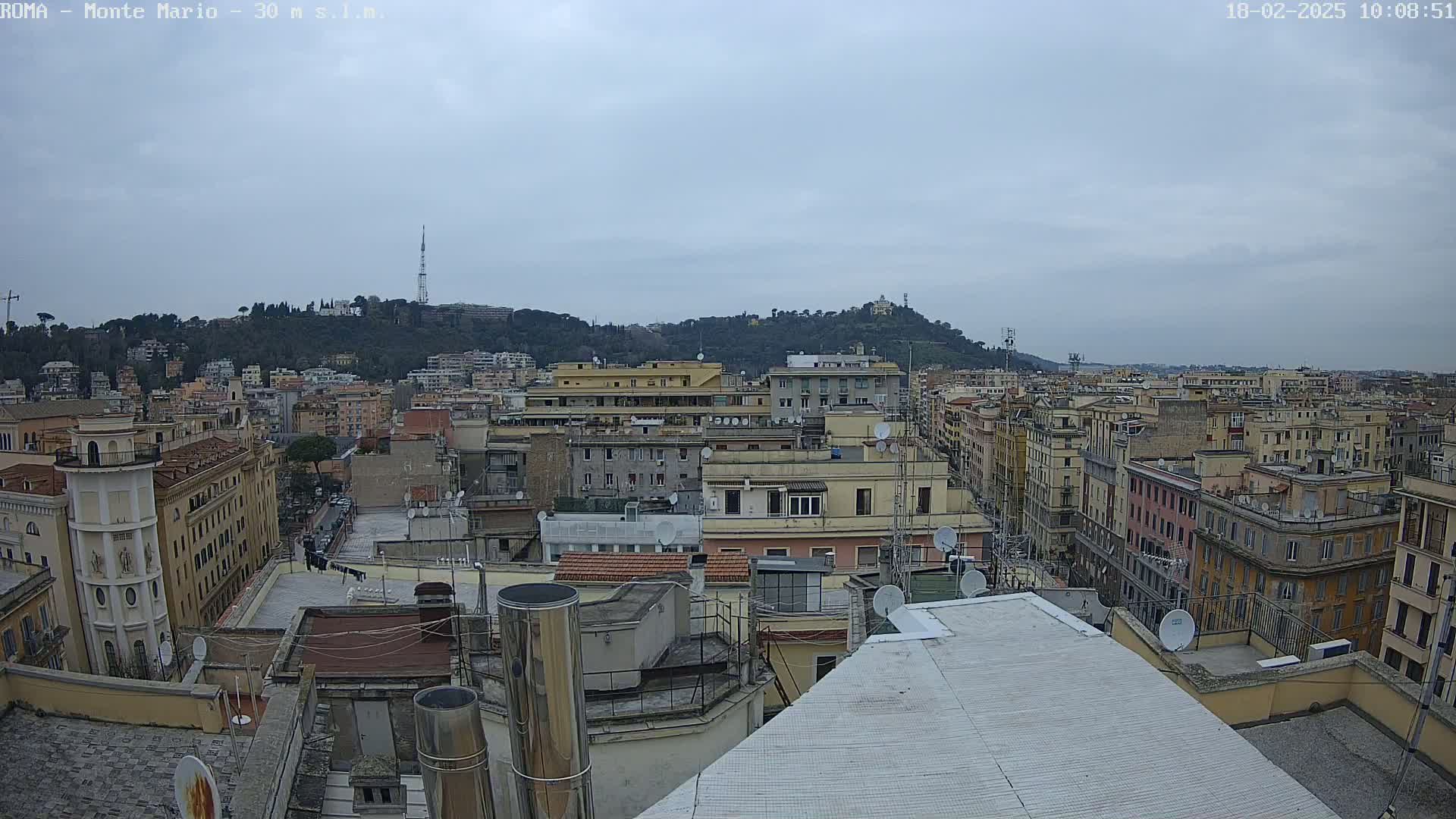 A high-angle view shows a cityscape of closely packed buildings under an overcast sky, with a hill visible in the background.