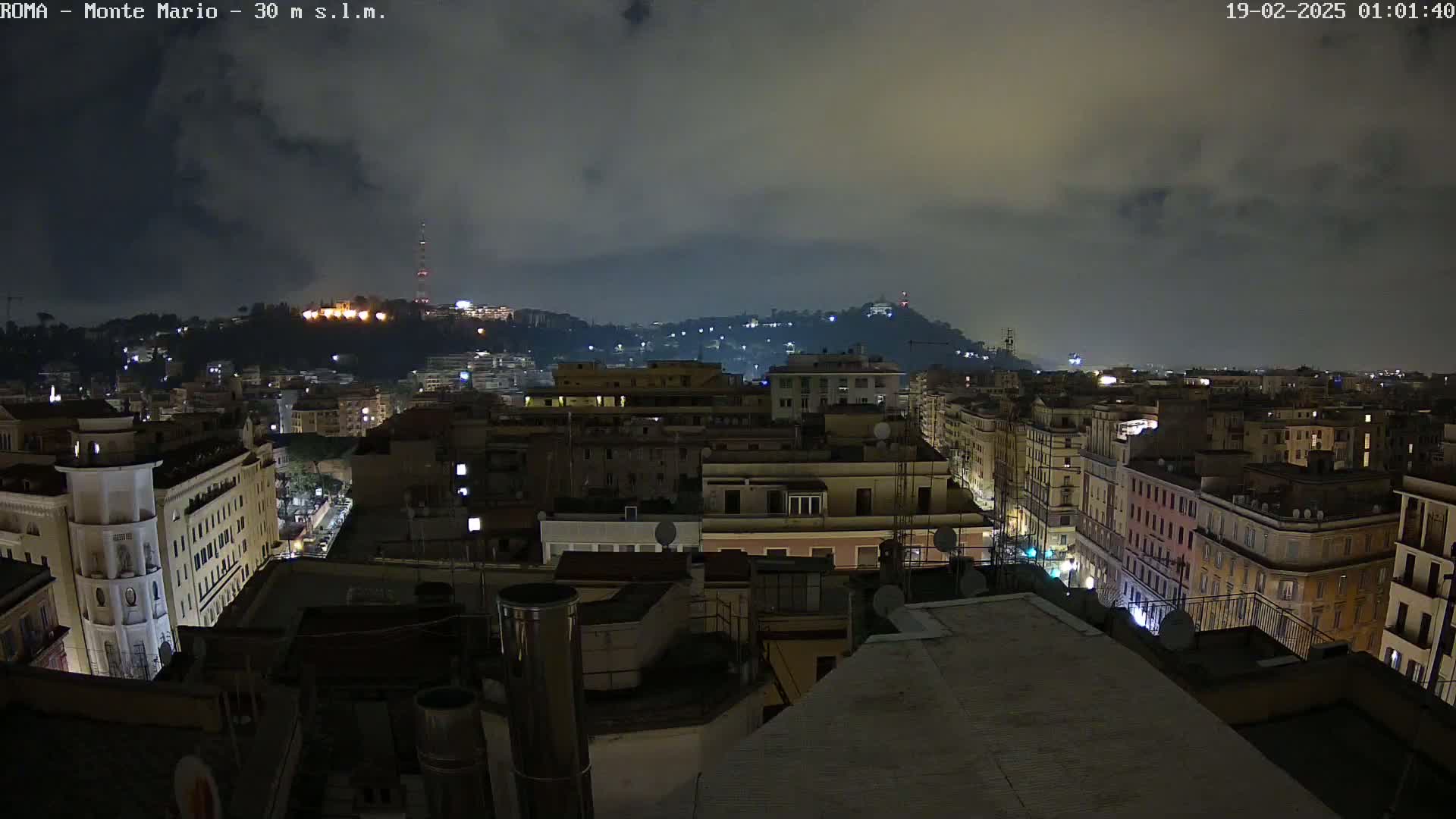 A nighttime cityscape view of a city with hills in the background under a cloudy sky.