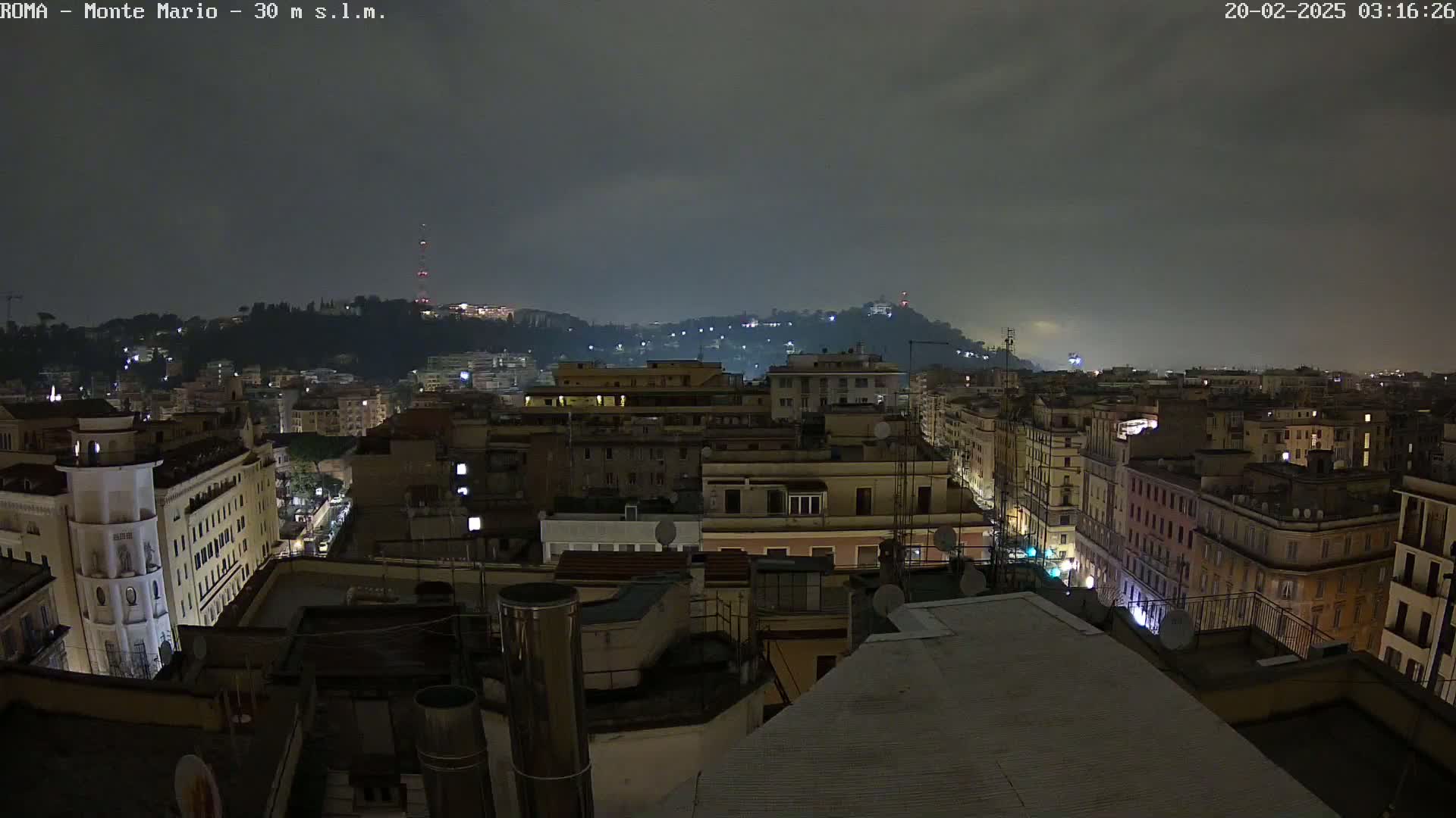 A nighttime cityscape under a cloudy sky shows numerous buildings with dimly lit windows, extending to a hill in the distance.
