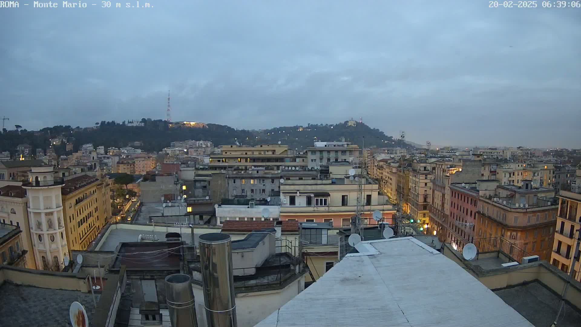 A rooftop view of a city at dusk under a cloudy sky, showing numerous buildings extending to a hill in the distance.