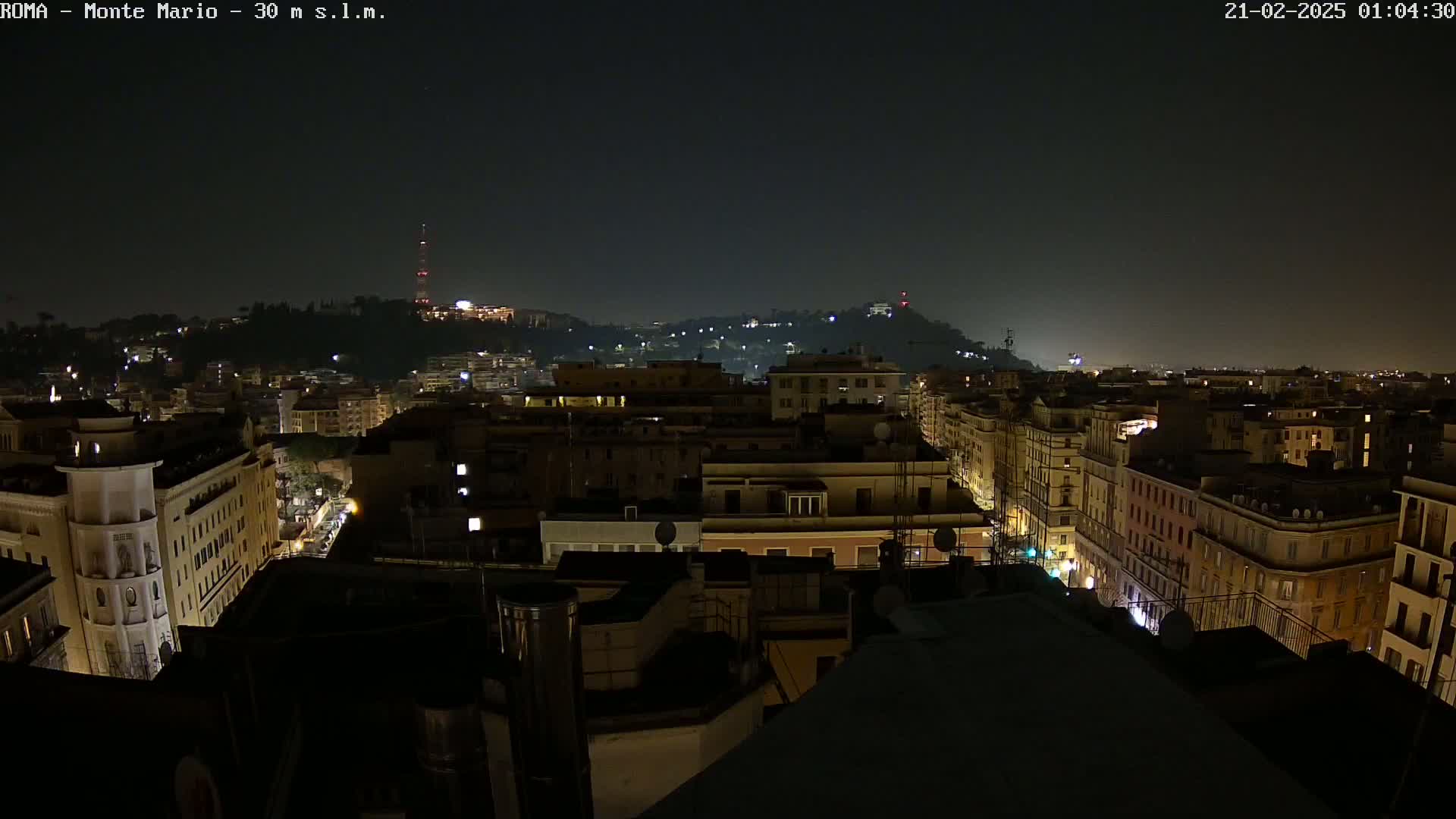 A nighttime cityscape view of illuminated buildings and streets, with a hill in the background under a clear, dark sky.