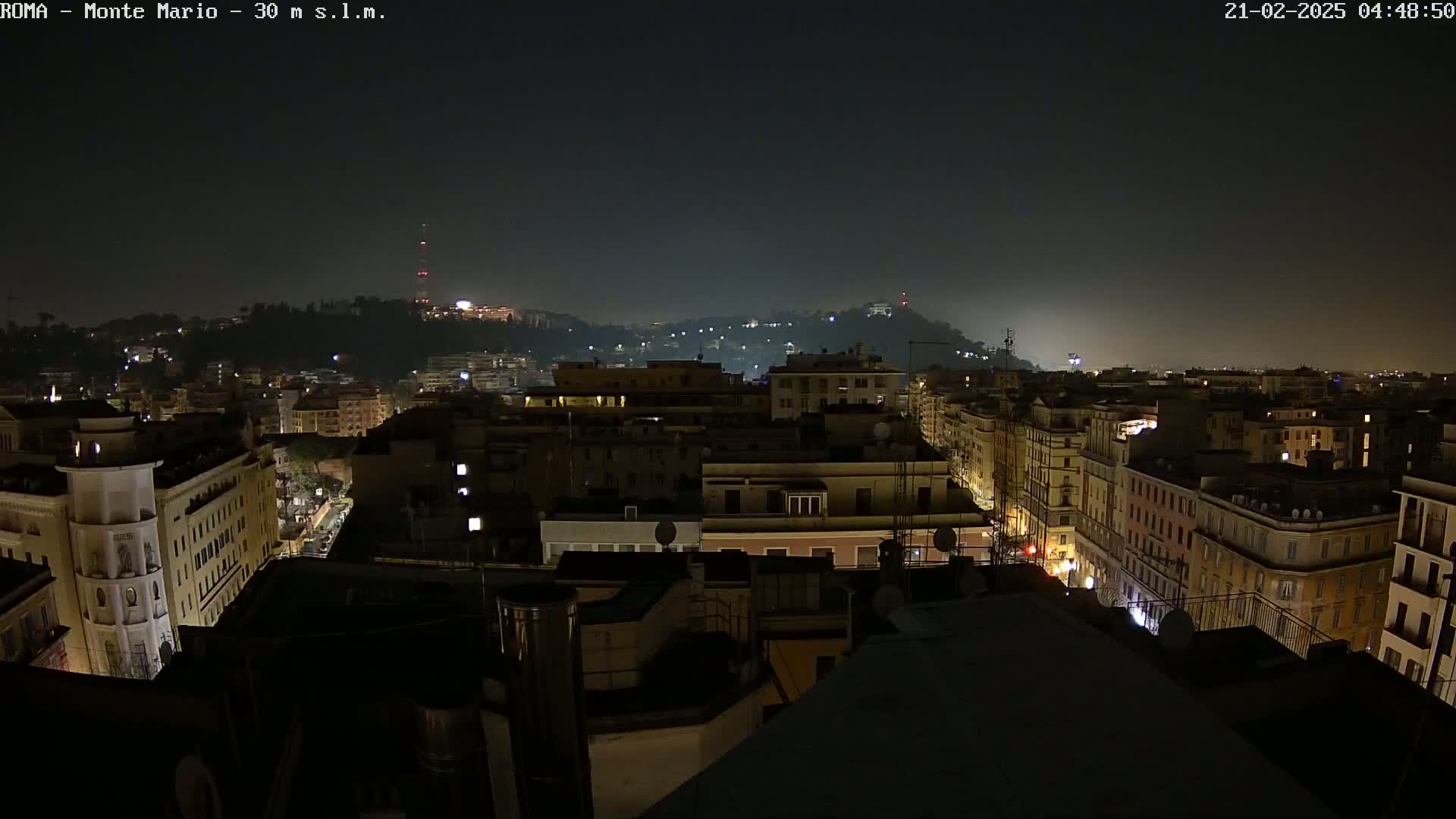 A nighttime aerial view of a city, showing a hilltop in the distance under a hazy sky.