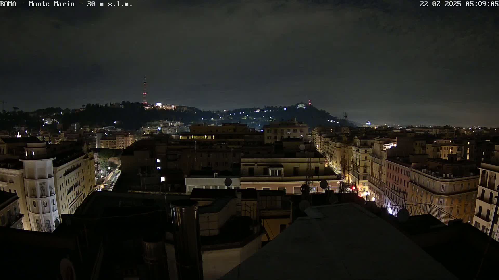 A nighttime view of a city with buildings illuminated, hills in the background under a dark, cloudy sky.