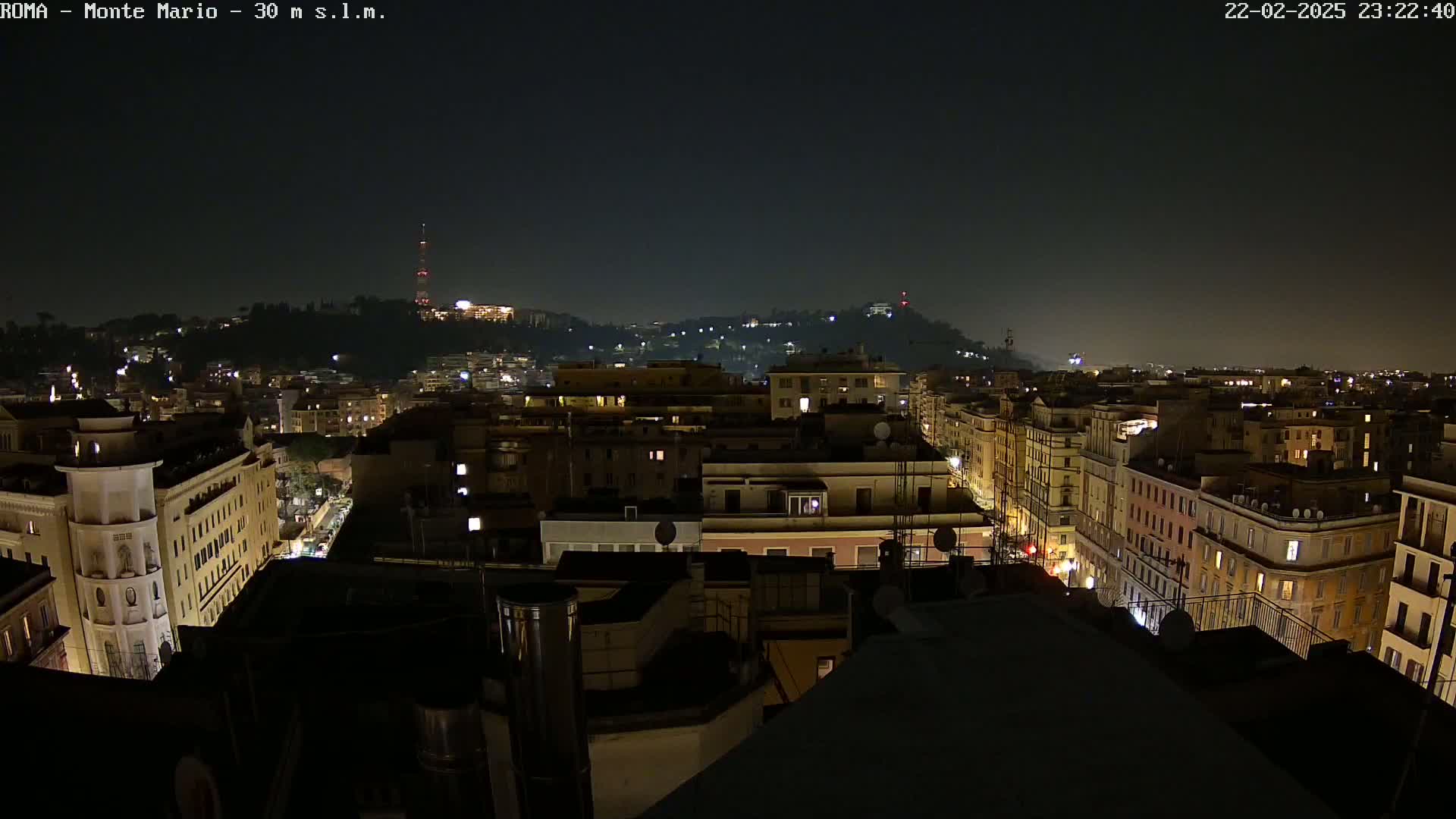 A nighttime aerial view of a city with numerous buildings illuminated, overlooking a hill in the distance under a clear, dark sky.