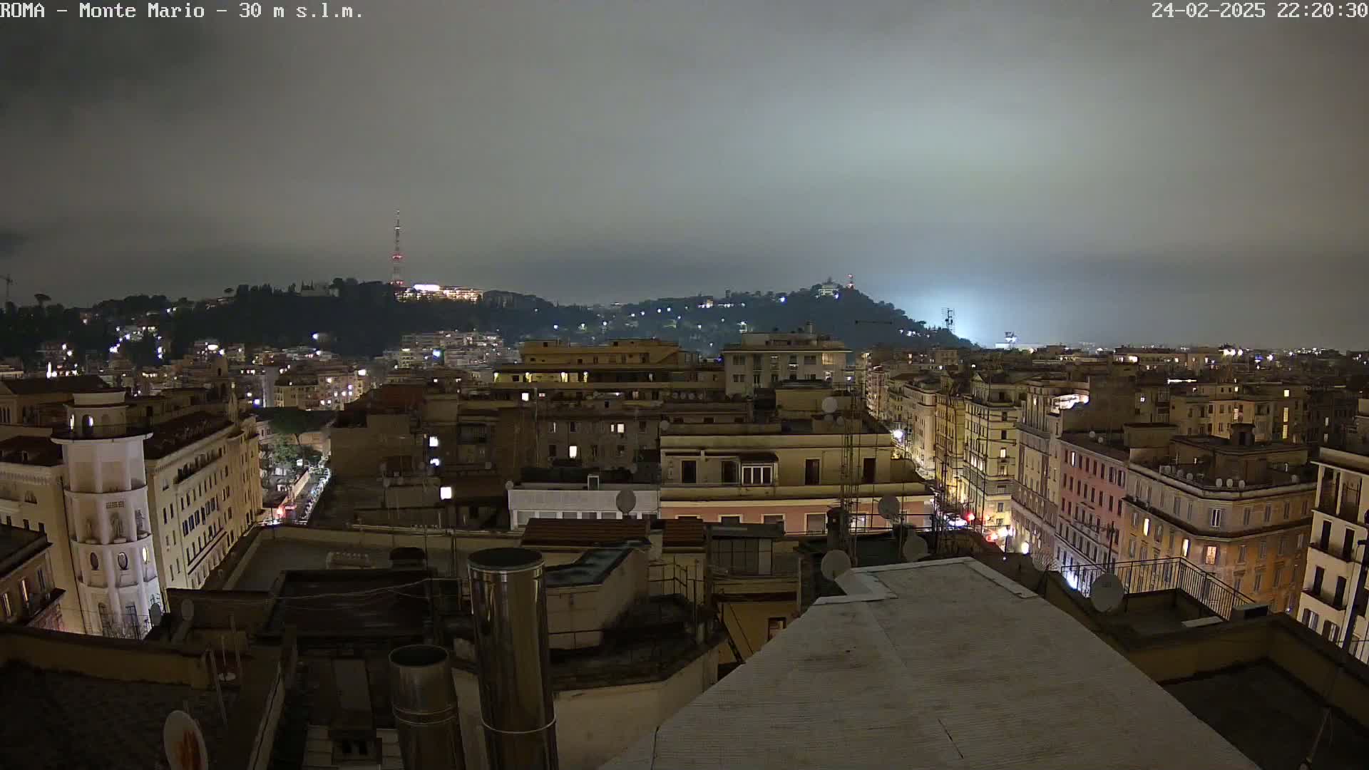 A nighttime view of a city under a cloudy sky shows numerous buildings illuminated with lights, extending to a hill in the distance.
