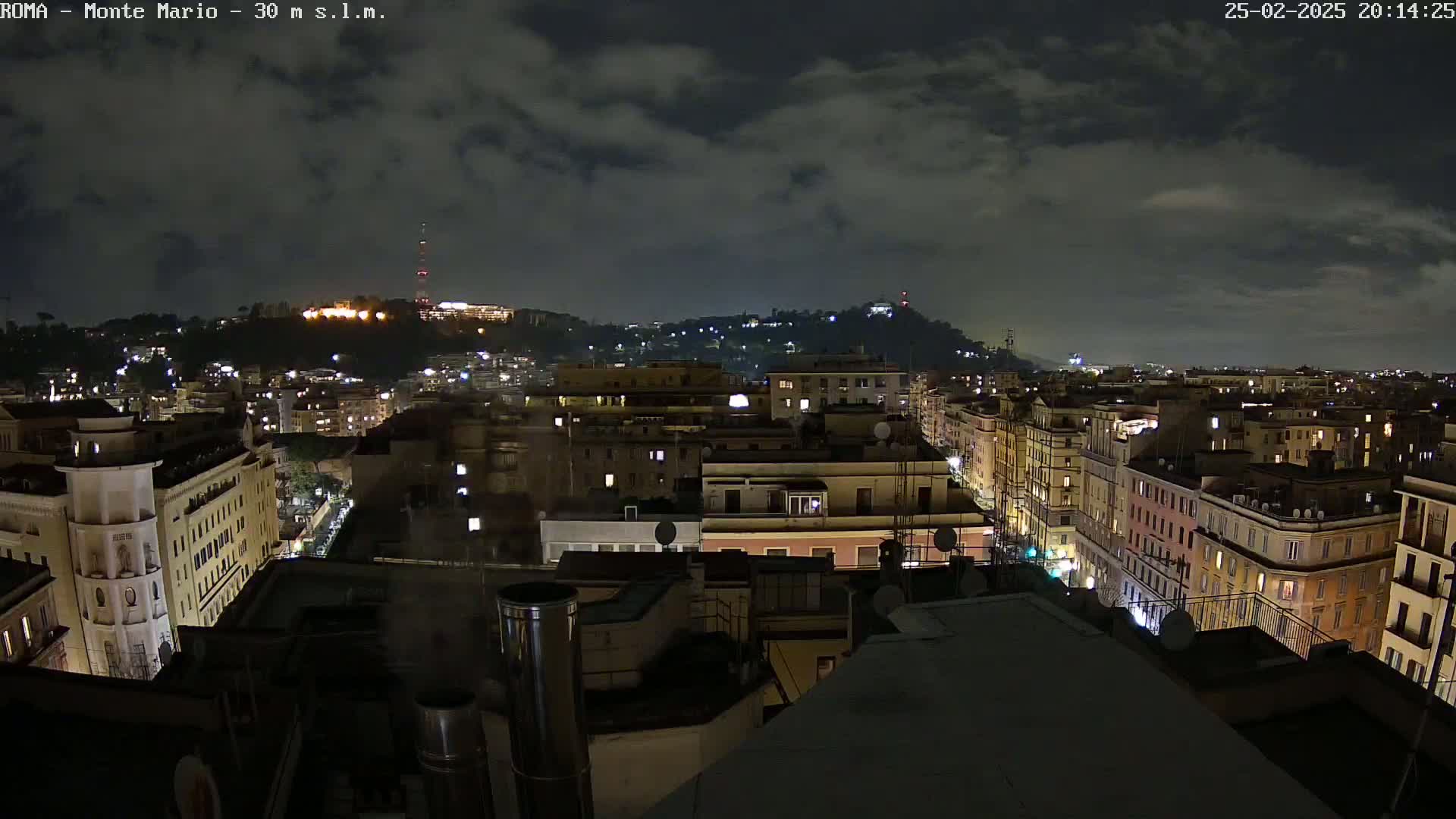 A nighttime view of a city, showing many buildings with lights on under a partly cloudy sky.