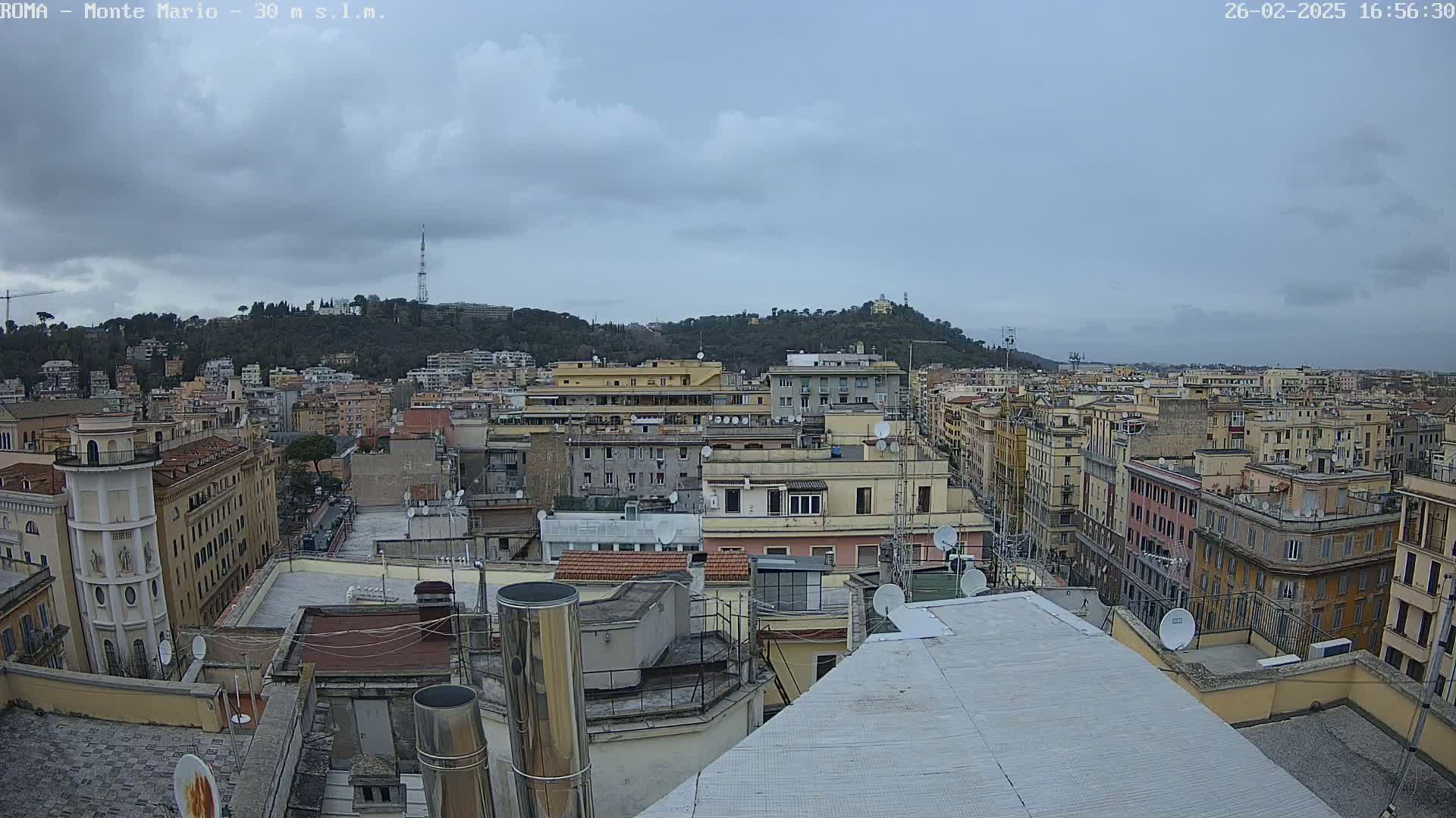 A view of a city under a cloudy sky, showing rooftops, buildings, and a hill in the distance.