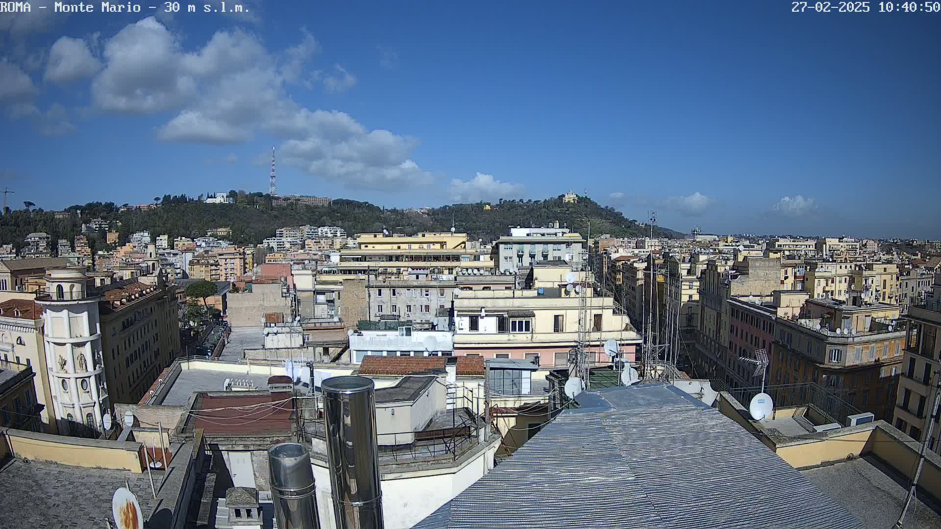 A mostly sunny day reveals a cityscape of closely packed buildings, with a hill topped by trees and structures visible in the background.