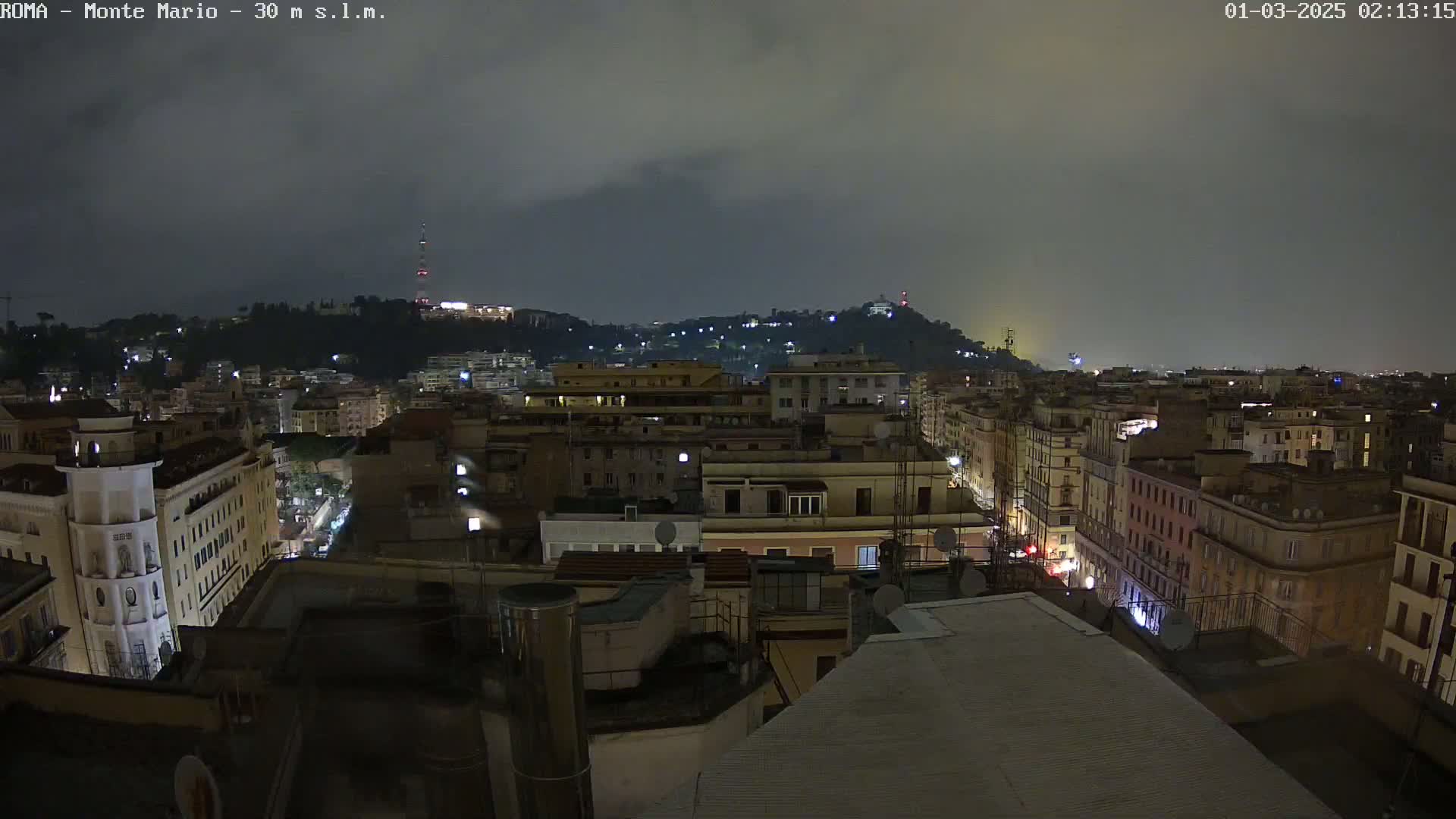 A nighttime cityscape view shows a hilly area with buildings and streetlights under a cloudy sky.