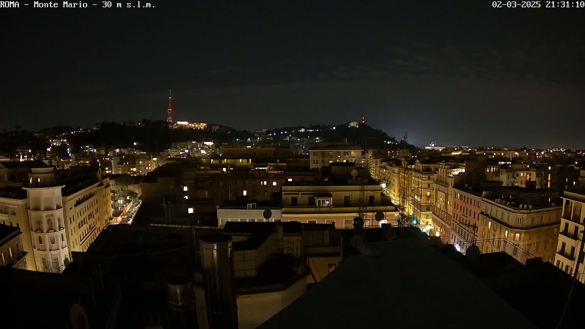 A nighttime cityscape of a city with illuminated buildings, stretching towards a hilltop in the distance under a dark, mostly clear sky.