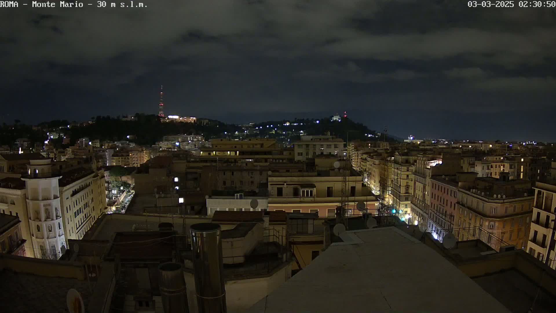 A nighttime aerial view of a city, under a cloudy sky, shows many buildings illuminated with lights, extending up a hill to a distant tower.