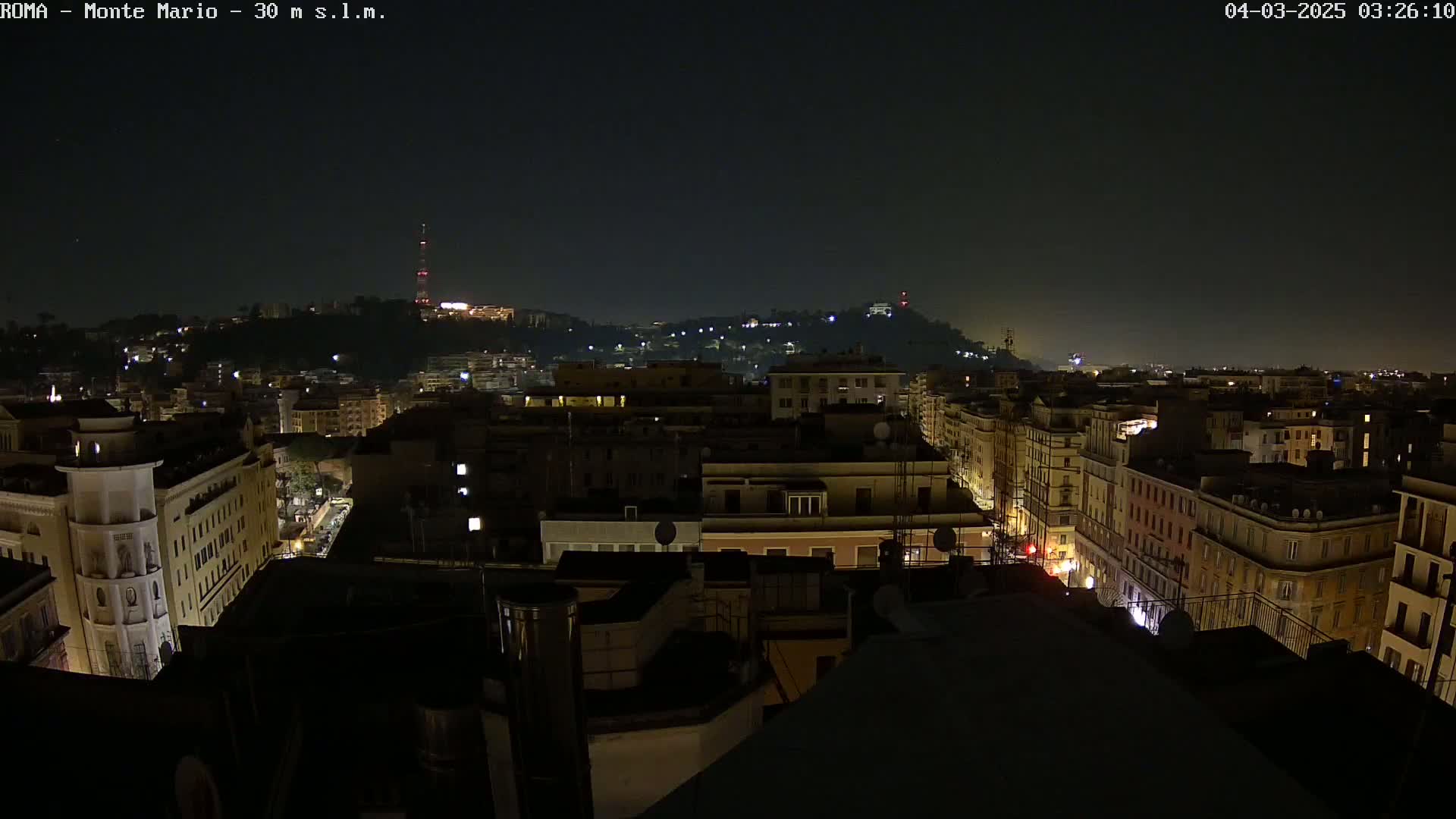 A nighttime, high-angle view of a city with illuminated buildings and streets, extending to hills in the distance under a clear, dark sky.