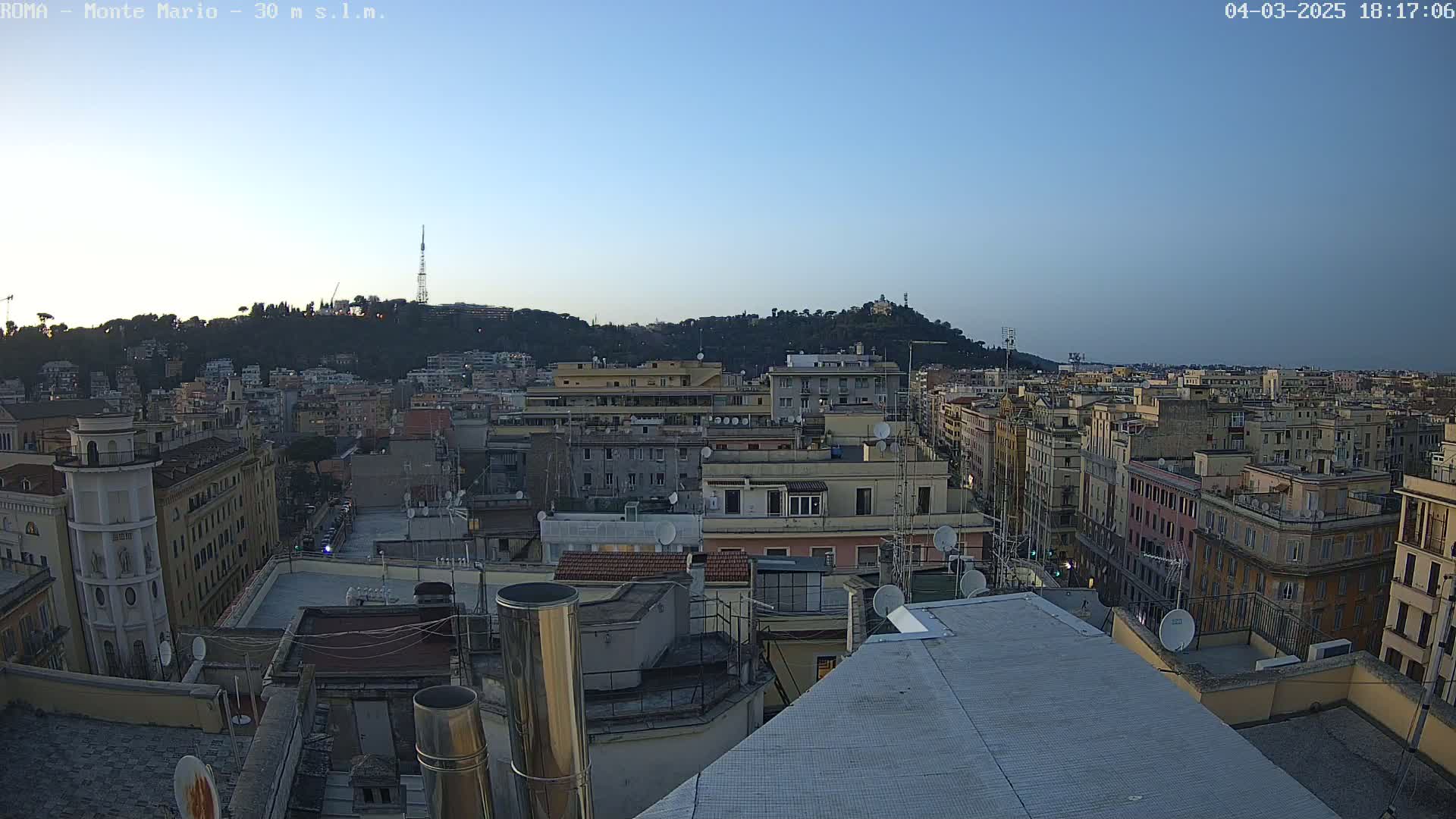 A clear twilight sky overlooks a cityscape of densely packed buildings, with a hill covered in trees visible in the background.
