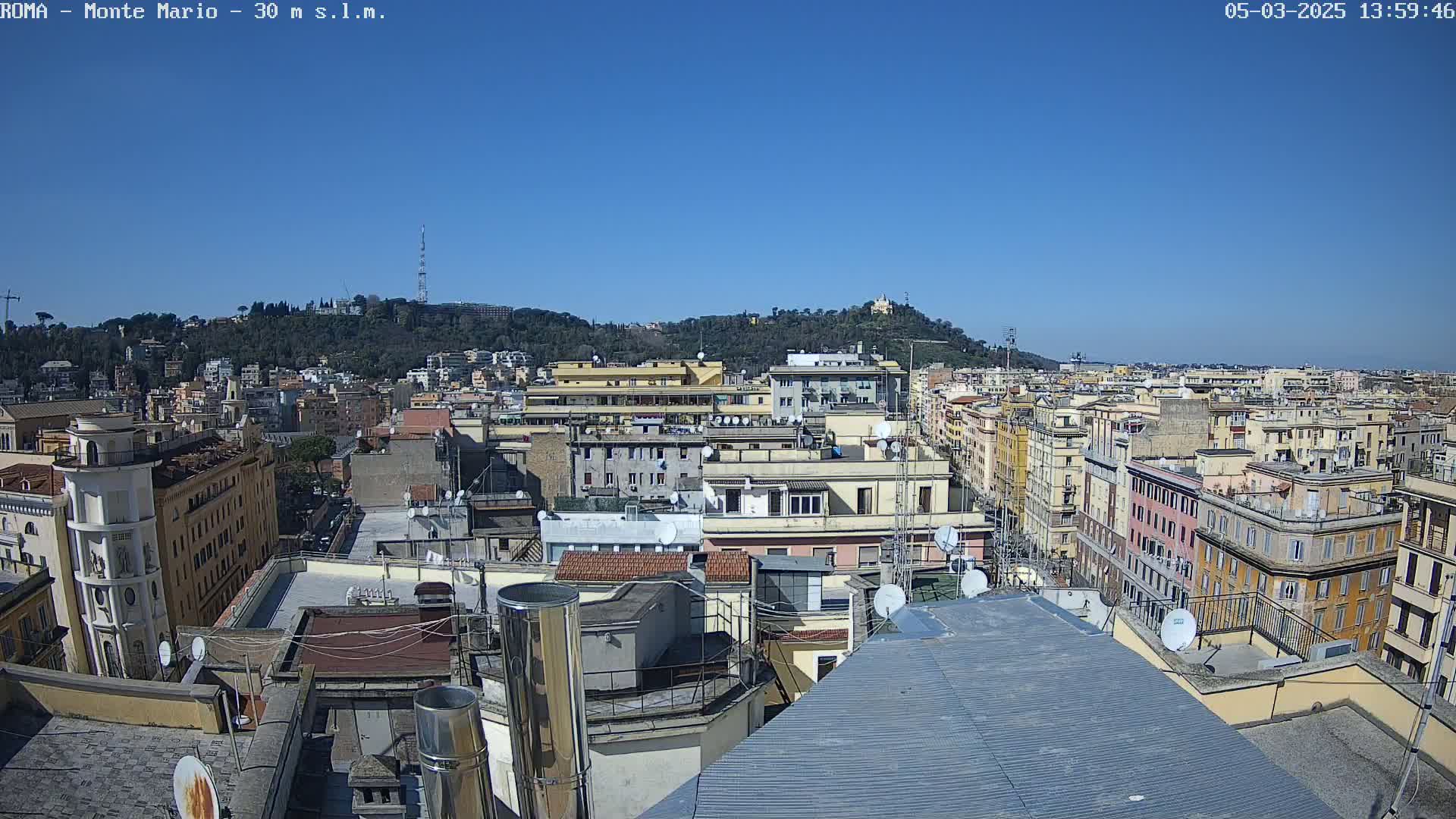 A high-angle view of a dense cityscape under a clear blue sky shows many multi-story buildings with rooftops and satellite dishes, extending to a tree-covered hill in the distance.