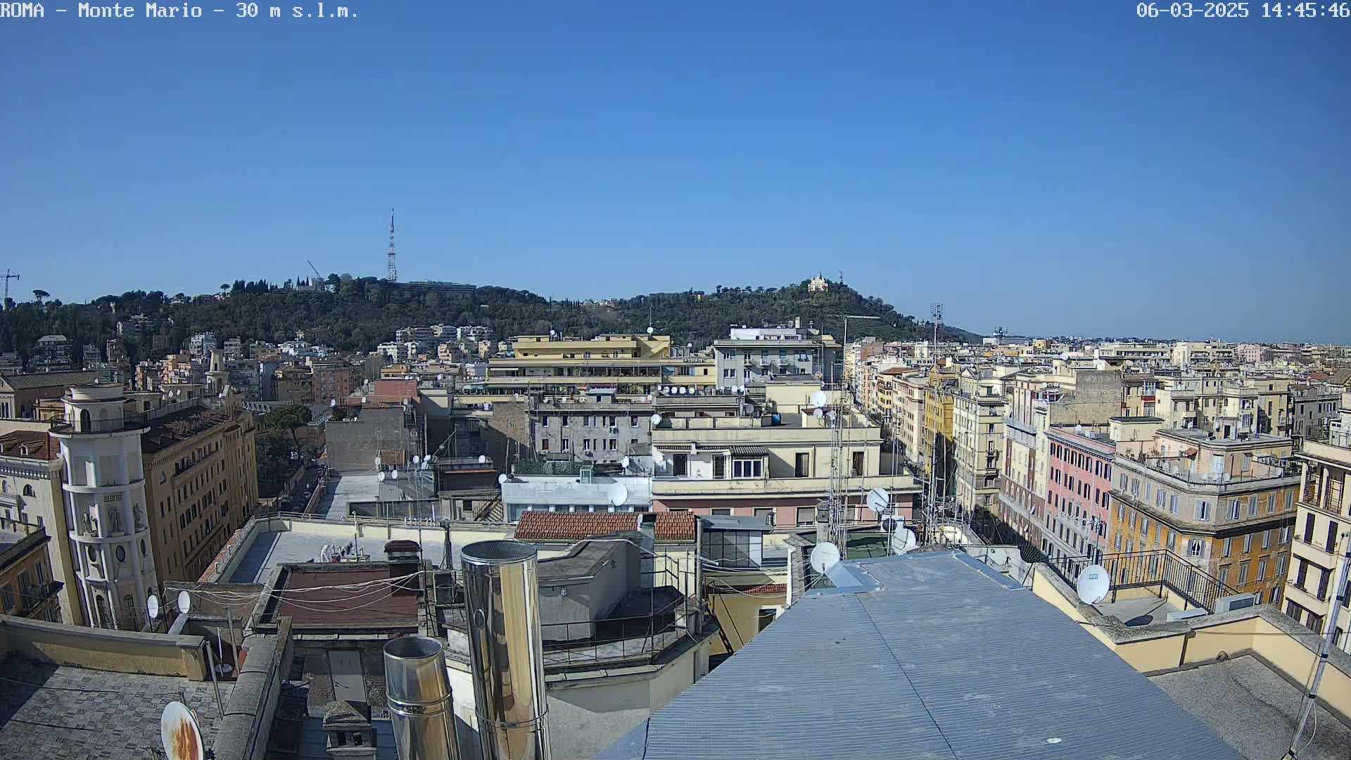 A sunny cityscape view from a rooftop shows numerous buildings, including multi-story apartment buildings and smaller structures, with a hill covered in trees in the background.