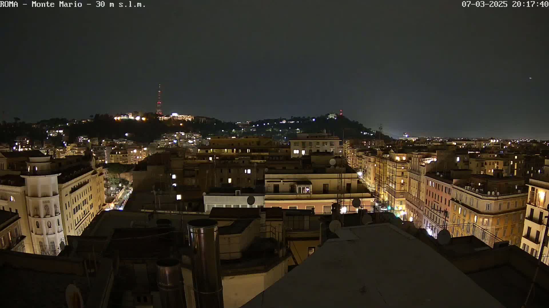 A nighttime view of a city, illuminated with lights from buildings and streetlights, shows a hill in the background under a dark, clear sky.