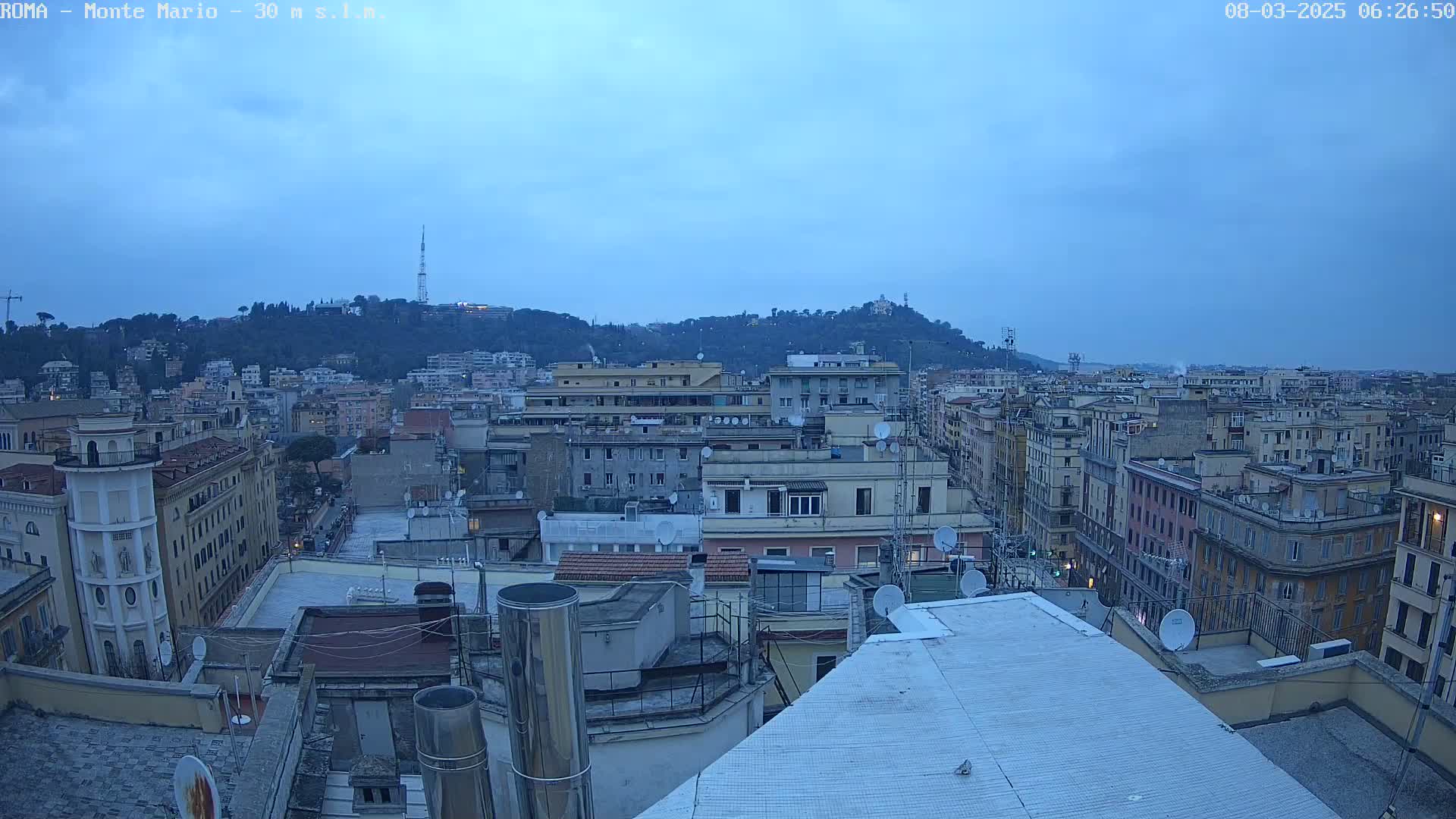 A rooftop view of a city under a twilight sky, showing many buildings and a hill in the distance.