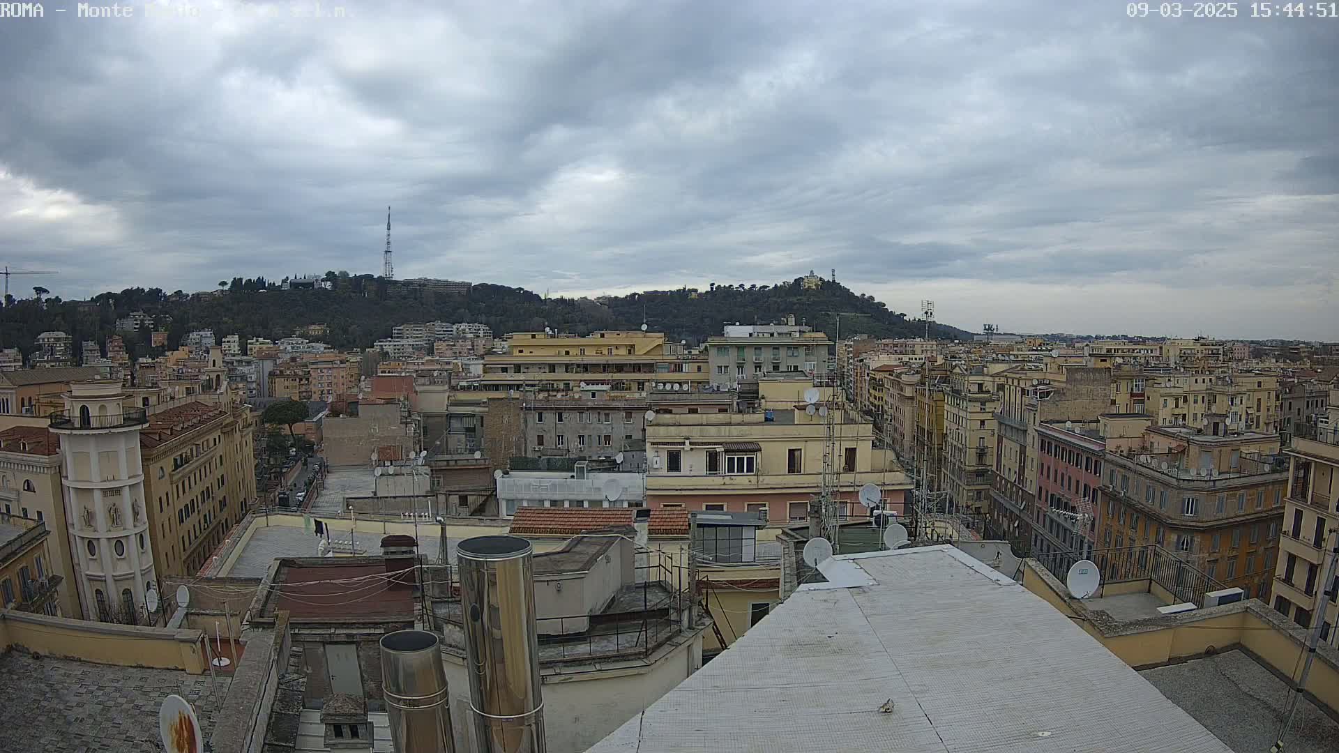 A view from a rooftop shows a city under a cloudy sky, with numerous buildings and a hill in the background.