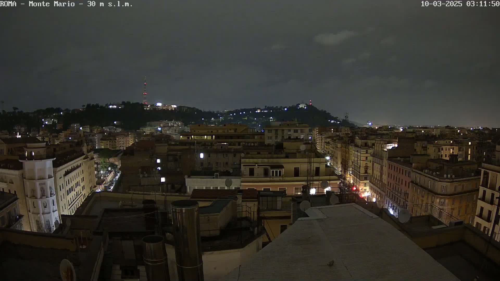 A nighttime view of a city with a hill in the background under a dark, cloudy sky.