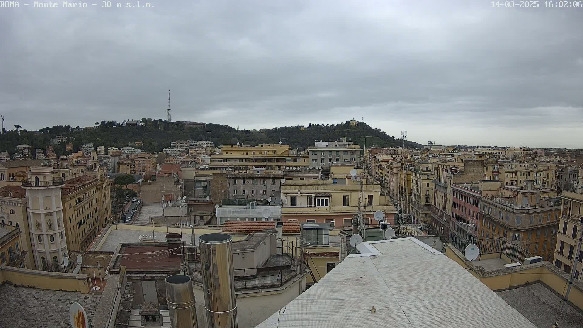 A cityscape of closely packed buildings under a cloudy sky, with a hill visible in the distance.