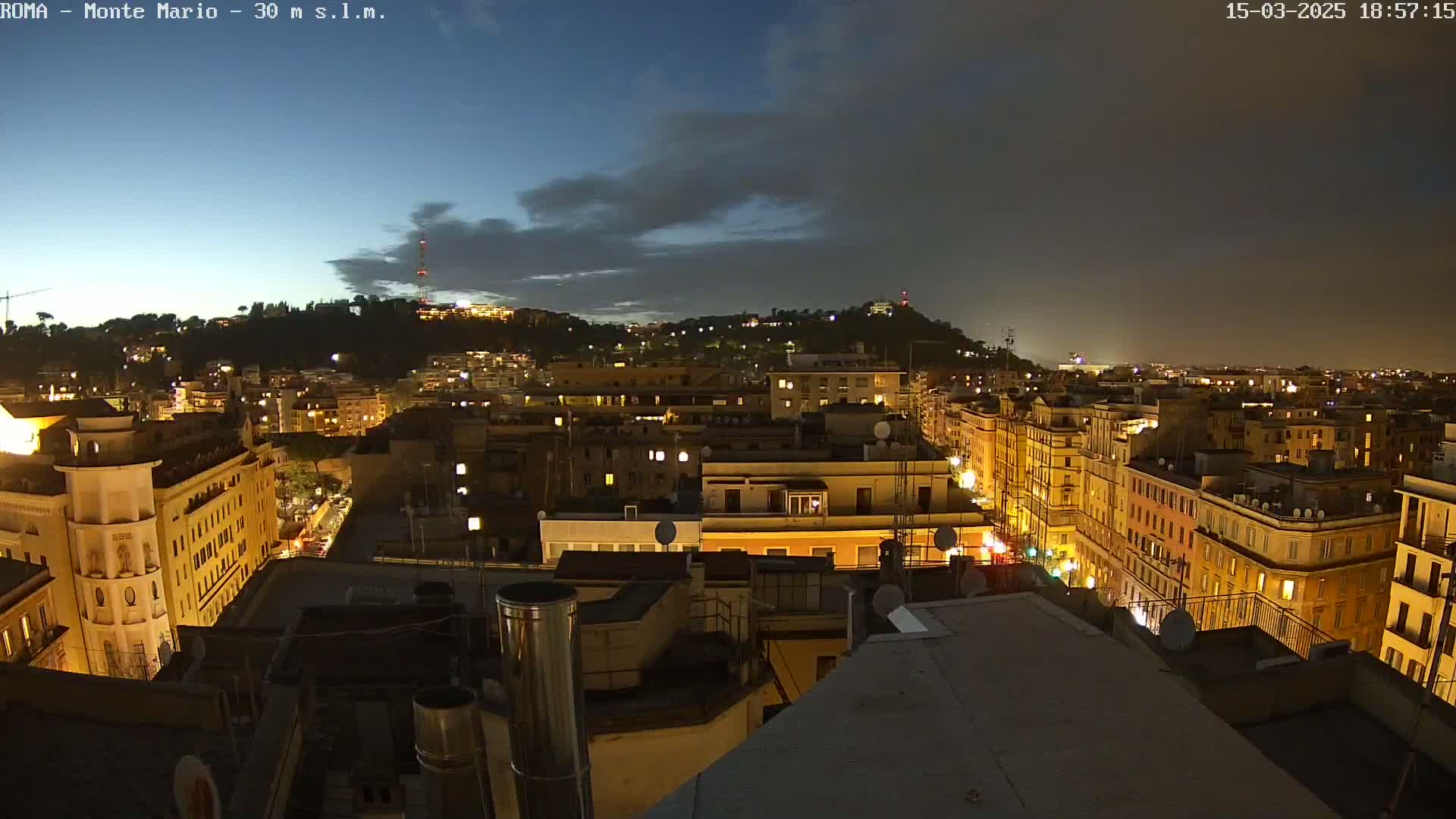 A nighttime cityscape of densely packed buildings illuminated by streetlights and window lights under a partly cloudy sky.