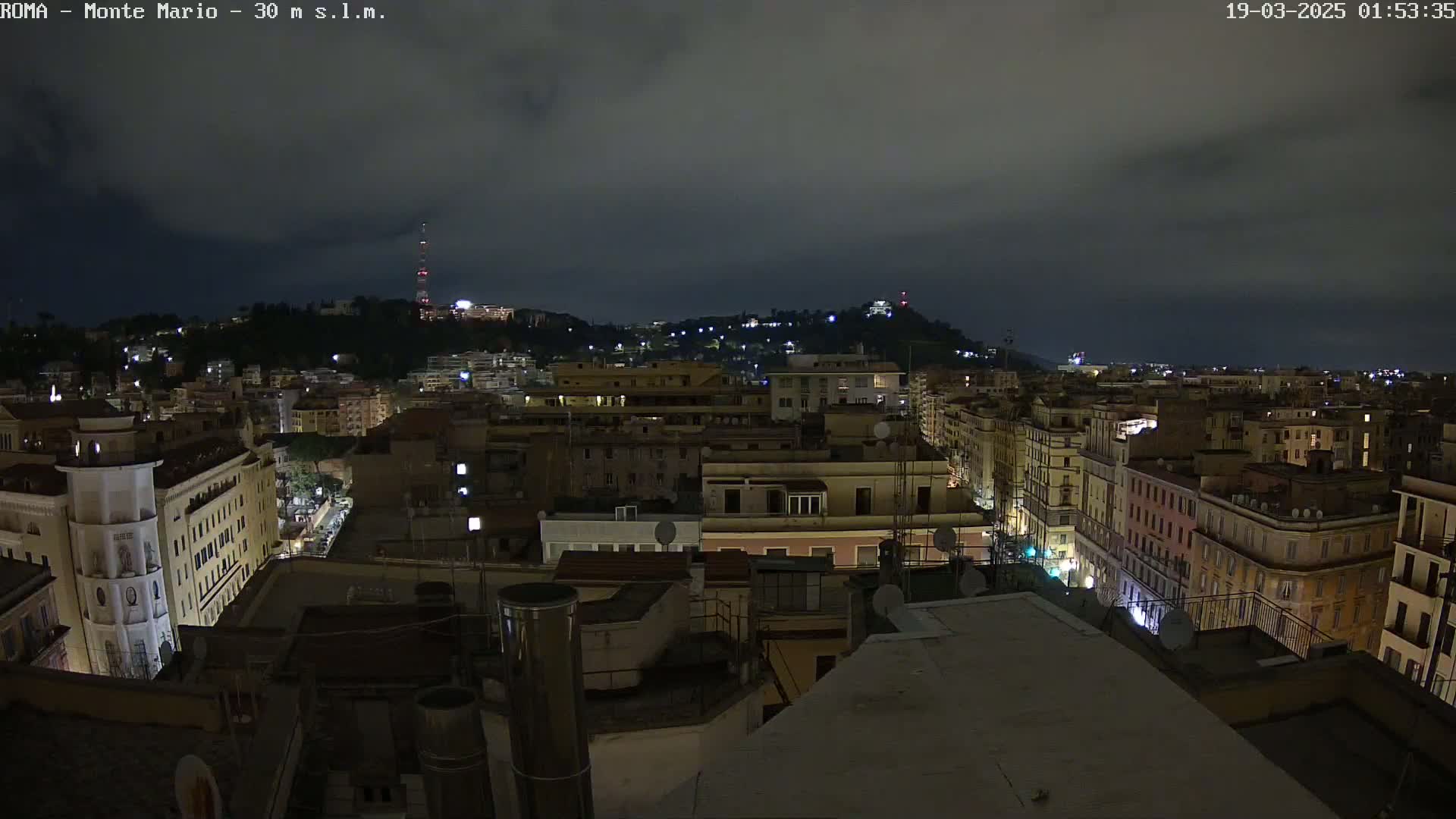 A nighttime view of a city, under a cloudy sky, shows numerous buildings illuminated with streetlights.