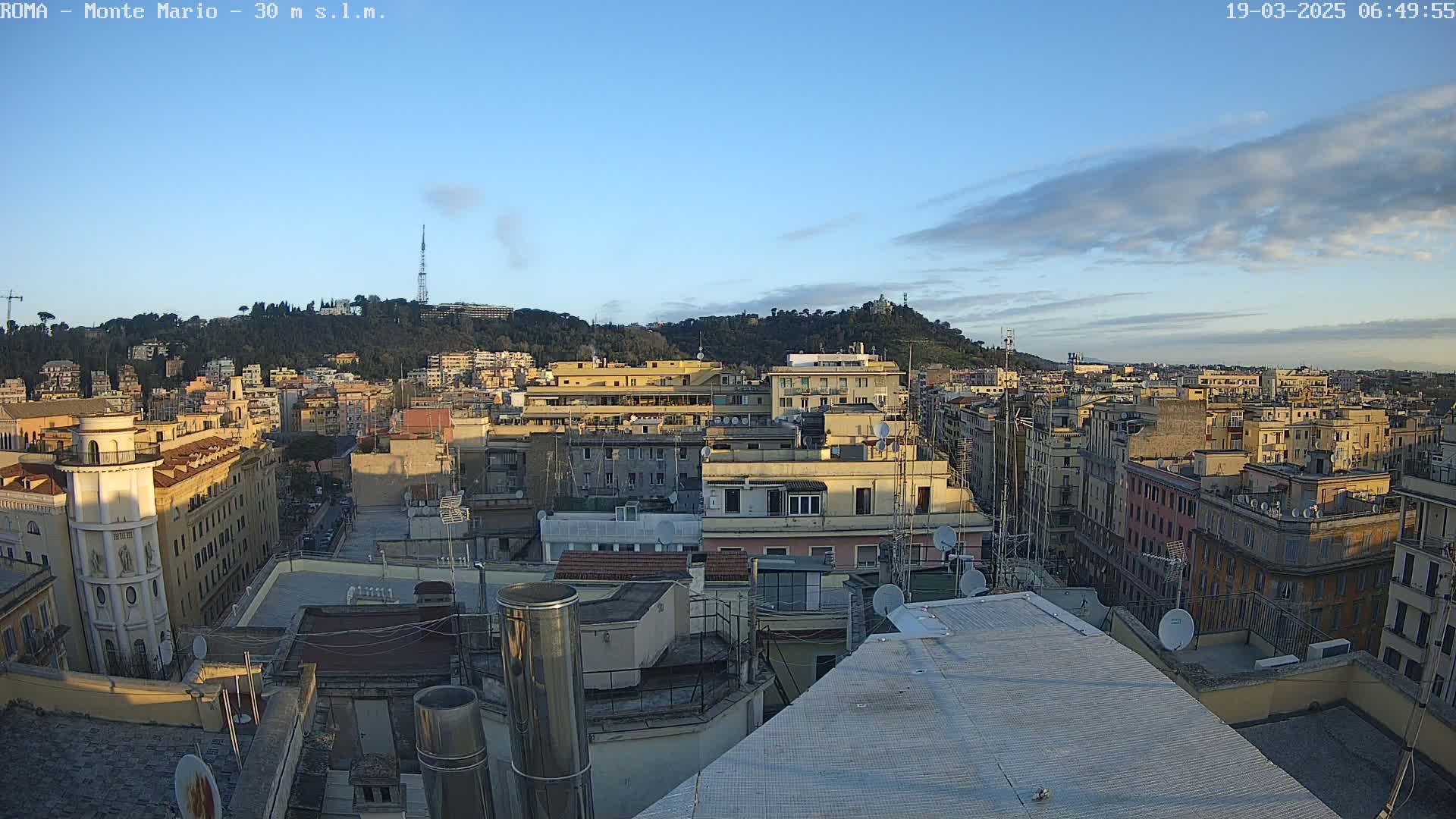 A mostly clear, early morning sky is over a cityscape of closely packed buildings, many topped with satellite dishes, with a tree-covered hill in the background.