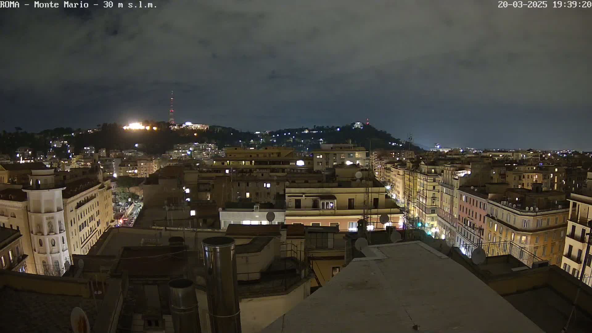 A nighttime view of a city, showing a hilly cityscape with illuminated buildings under a cloudy sky.
