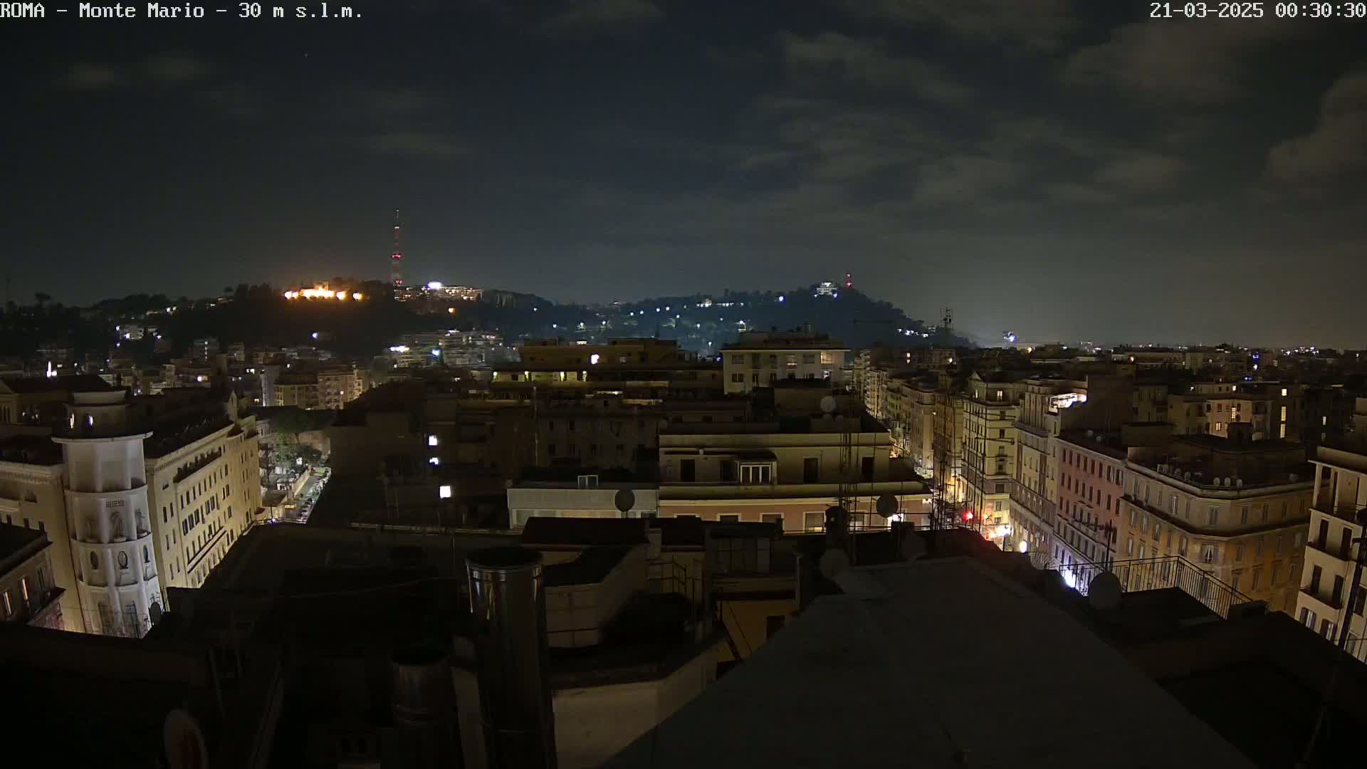 A nighttime view of a city, under a partly cloudy sky, showing a cityscape illuminated by lights from buildings and streets.