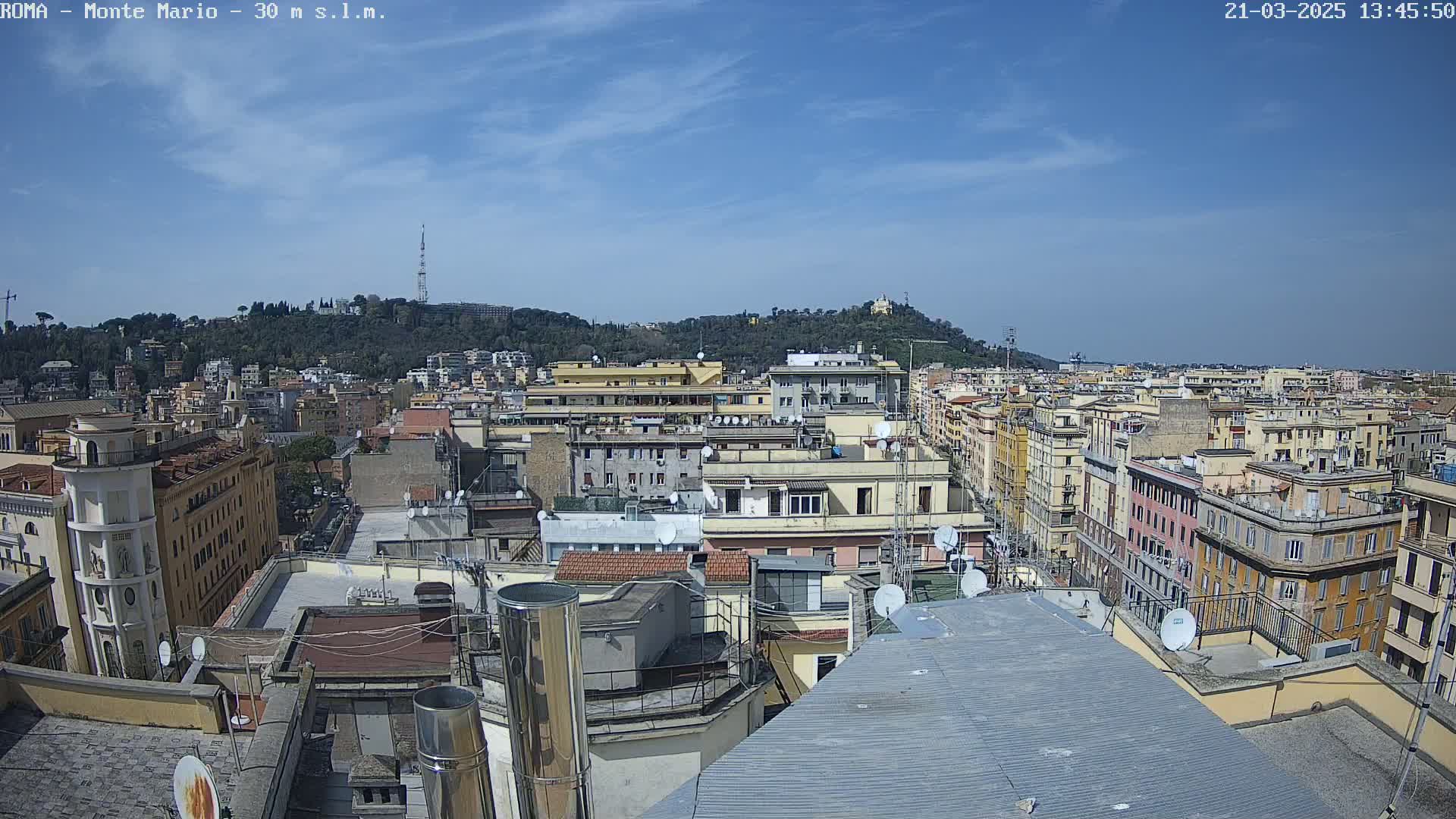 A cityscape of densely packed buildings under a mostly clear, sunny sky, with a hill covered in vegetation visible in the background.