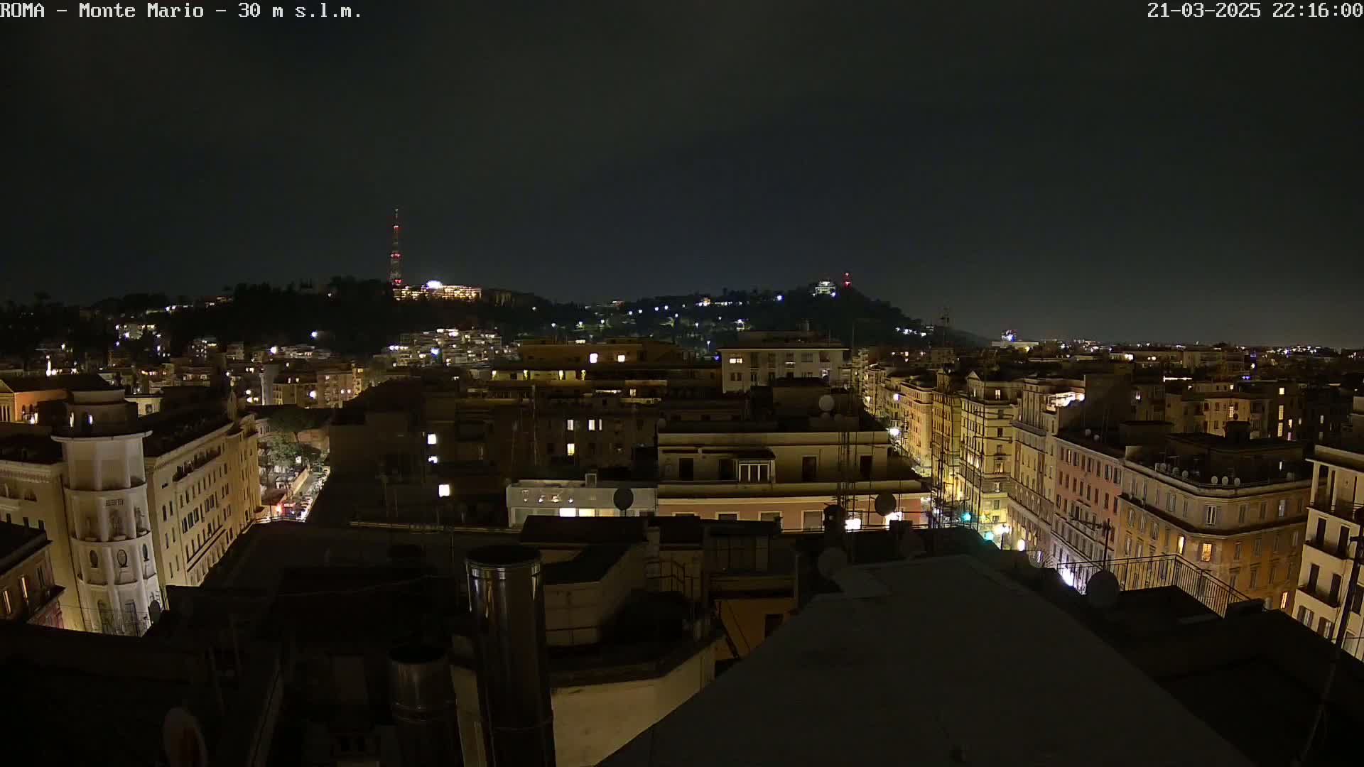 A nighttime cityscape view shows a hilly area with illuminated buildings, streets, and a distant tower under a dark sky.