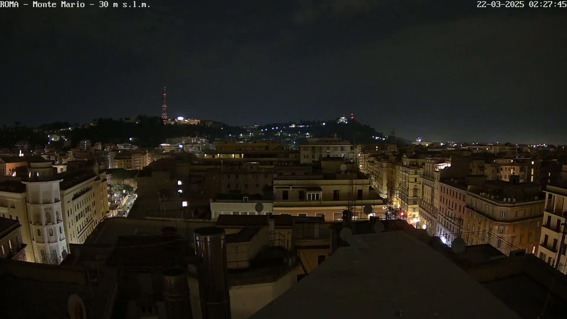 A nighttime cityscape shows a hilltop in the distance with a tall tower, and many buildings with windows illuminated below, under a dark, clear sky.