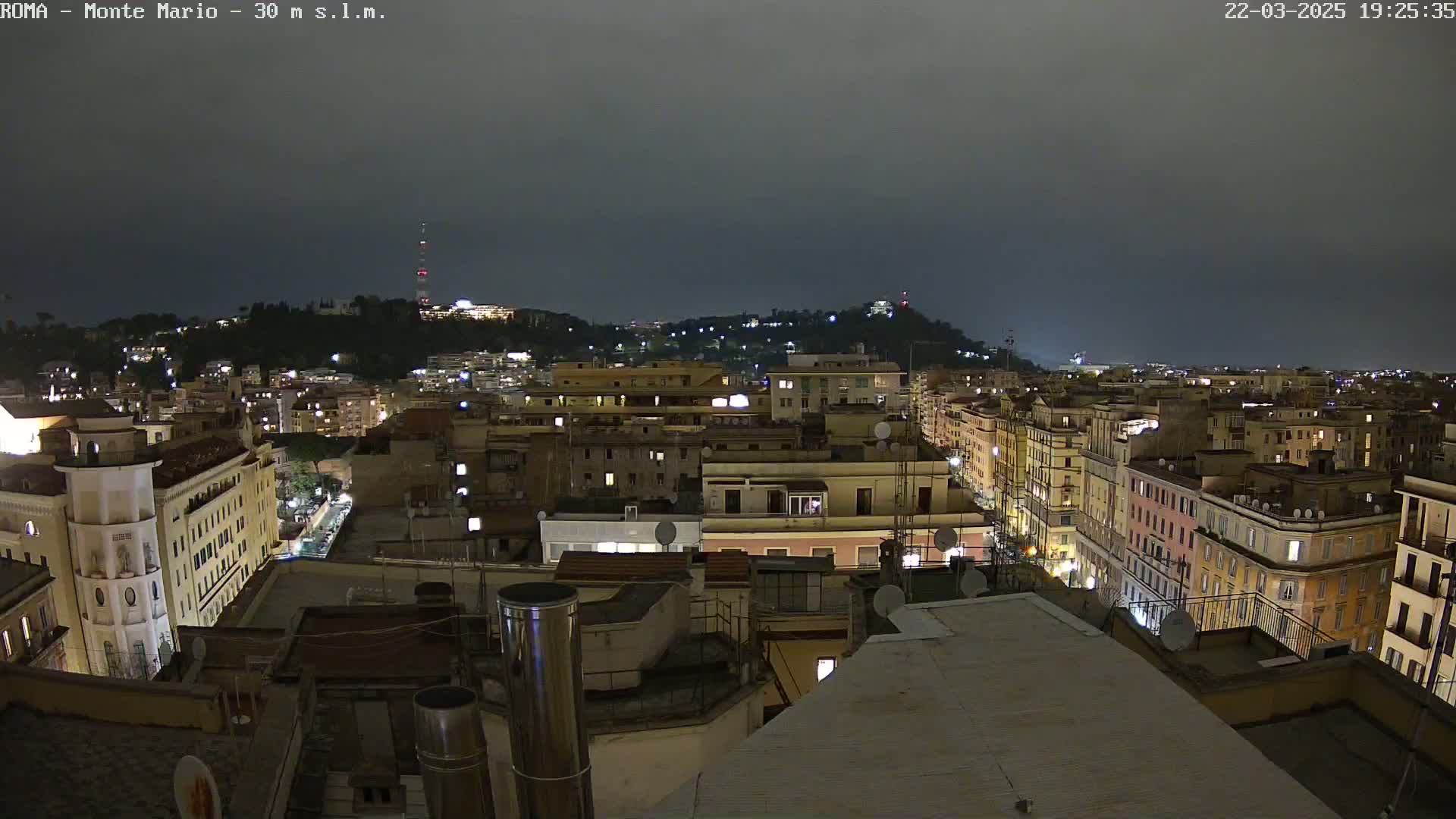 A nighttime view of a city, under a dark, cloudy sky, shows numerous buildings lit up with windows glowing, extending to a hill in the distance.