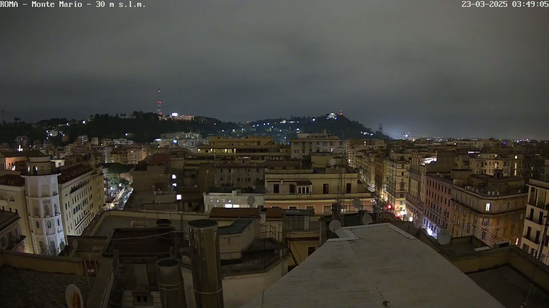A nighttime view of a city, under an overcast sky, shows numerous buildings illuminated against a hill in the distance.