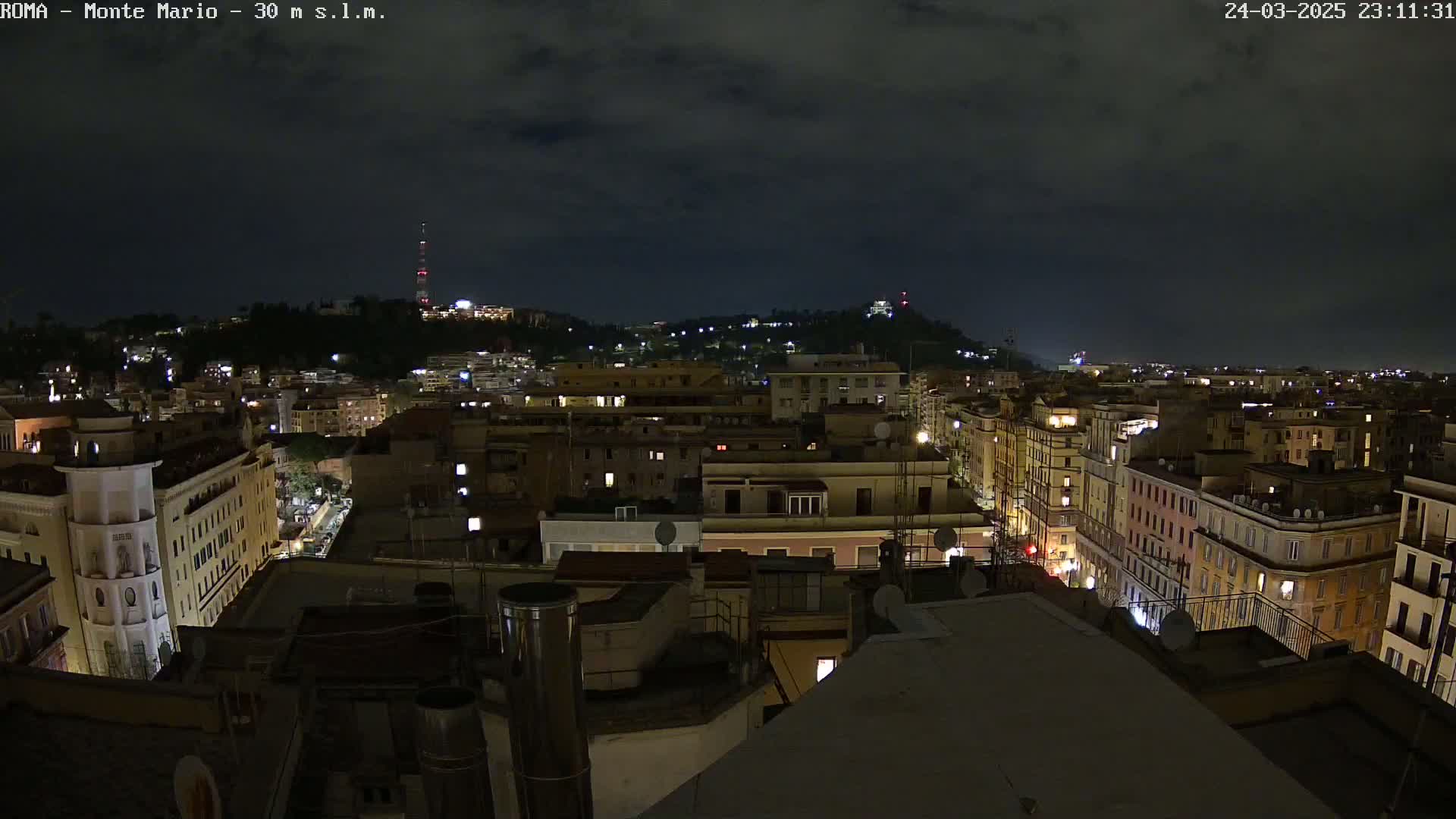 A nighttime view of a city, showing many buildings with lights on under a dark, cloudy sky.