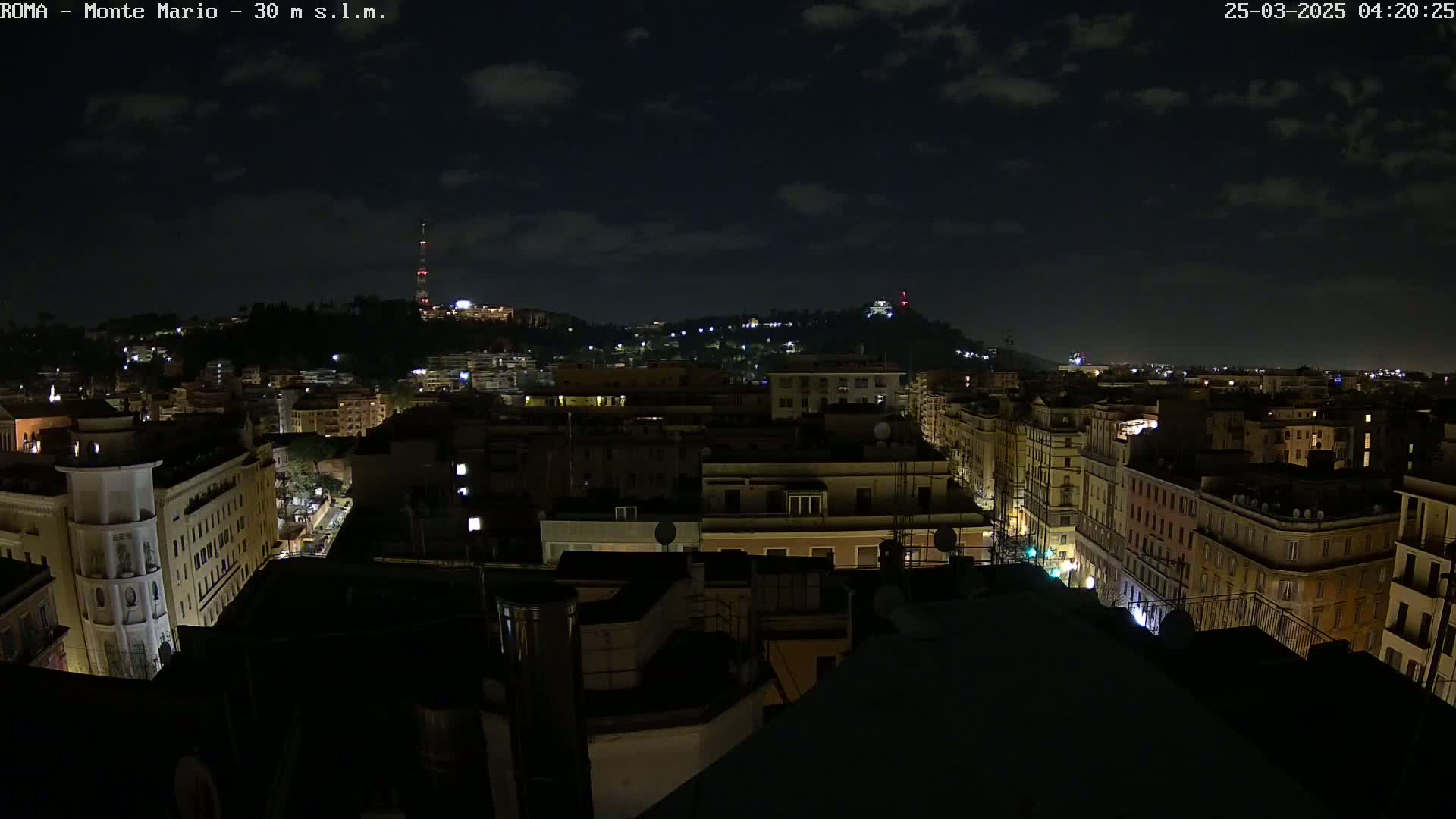 A nighttime view of a city, showing a hilly landscape with buildings lit up under a partly cloudy sky.