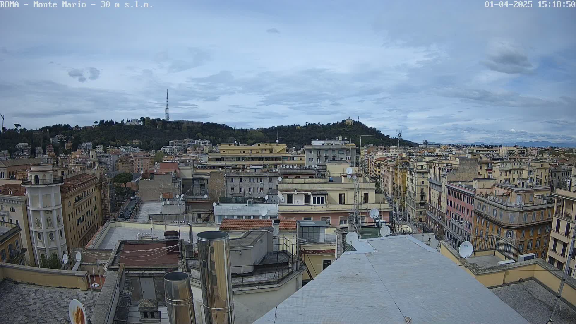 A high-angle view of a city under a mostly cloudy sky, showing numerous buildings and rooftops, with a hill in the background.