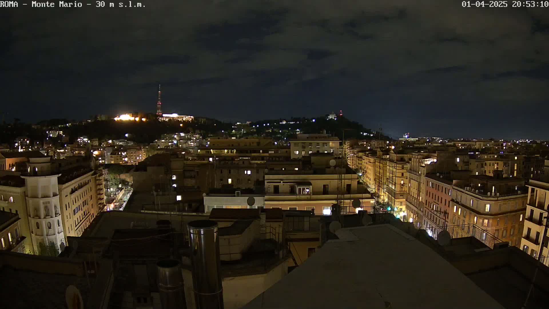 A nighttime view of a city with illuminated buildings and a hill in the background under a partly cloudy sky.