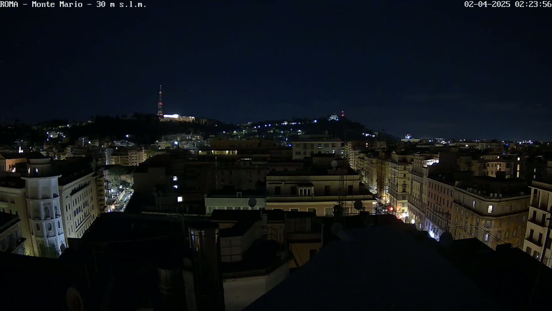A nighttime view of a city, showing illuminated buildings and streets extending to a hill in the distance under a clear, dark sky.