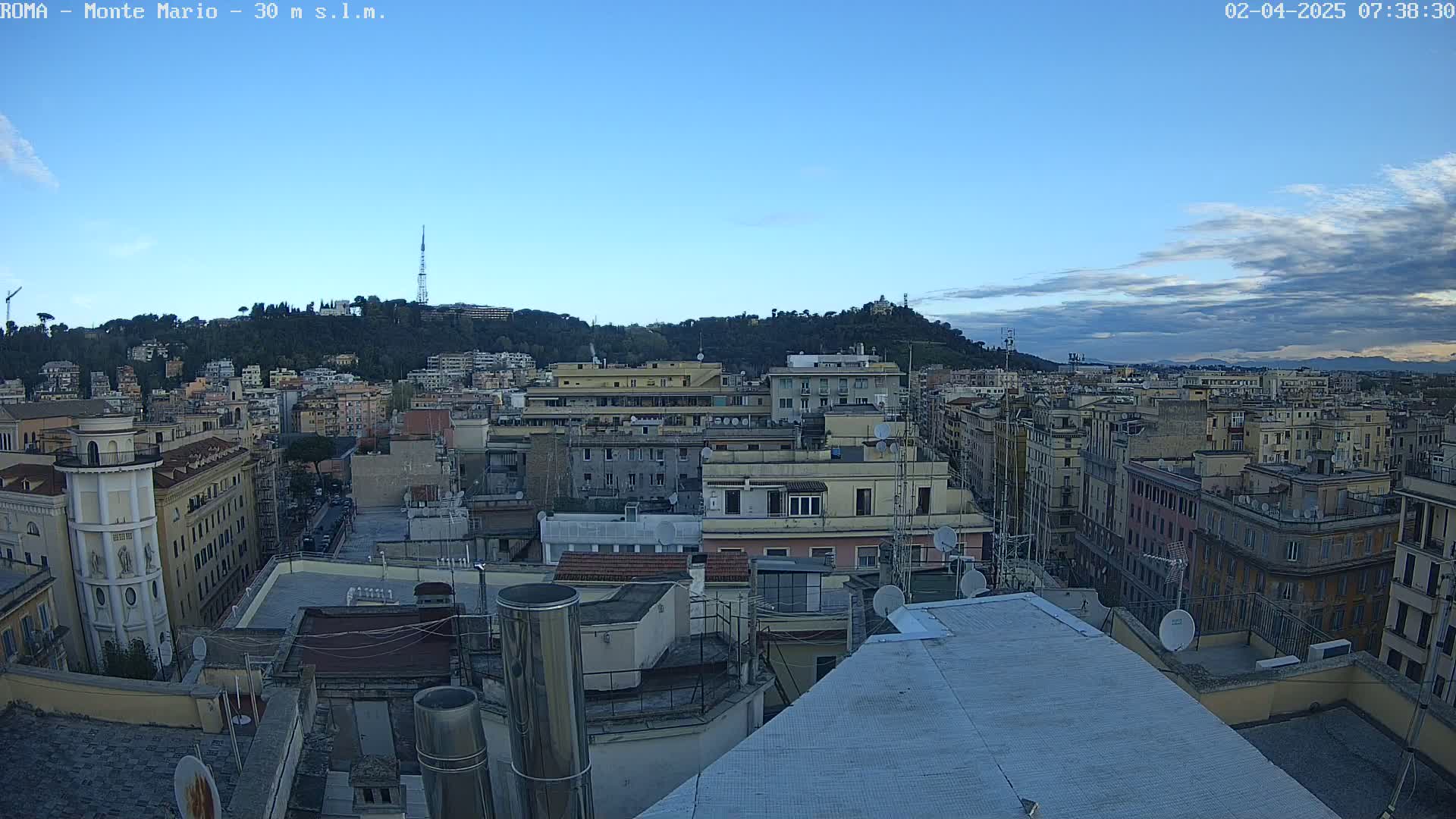 A high-angle view of a city landscape under a mostly clear, early morning sky shows numerous buildings and rooftops, with a hill in the background.