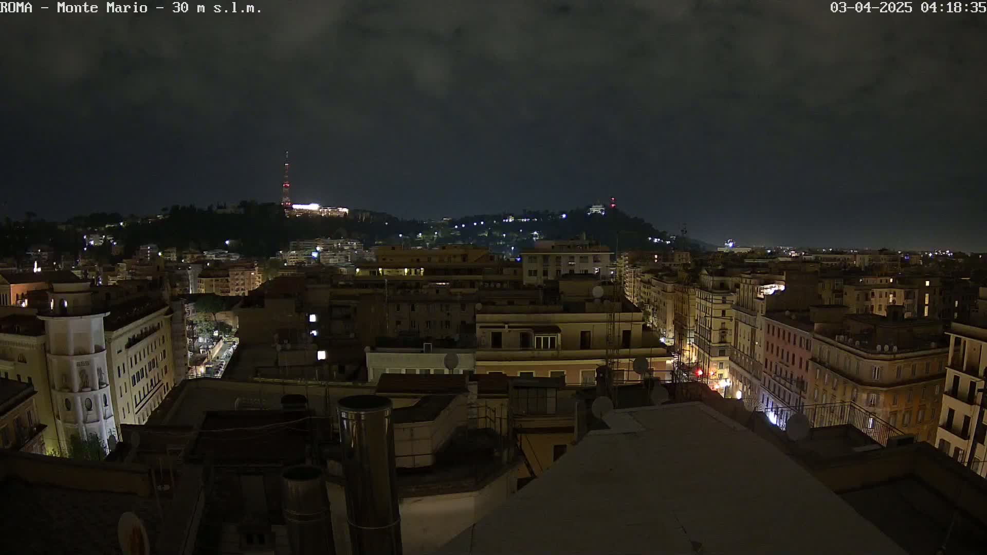 A nighttime view of a city, with a hilltop in the distance under a cloudy sky.